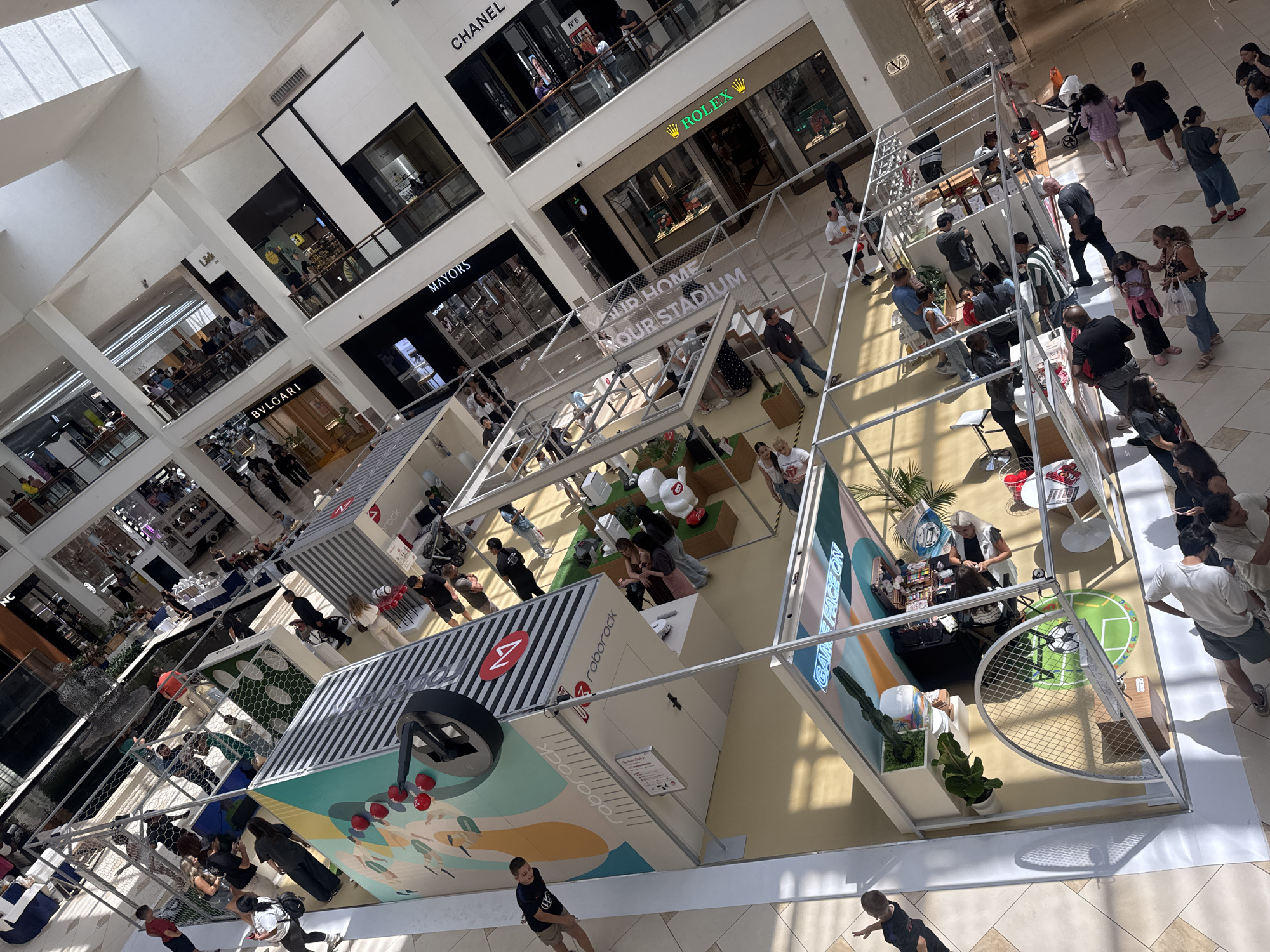 The image depicts a bustling interior scene of the Aventura Mall in Aventura, Florida, United States of America. The perspective is from above, looking down at a multi-level shopping mall with a central atrium. The architecture features a modern design with glass railings and metallic structural elements.
Several temporary exhibits or booths are set up on the ground floor. One prominent display features a large graphic of robots and a stylized wheel, with the text "roborock" visible. Another exhibit has the text "GAME FACE ON" and displays what appear to be gaming-related items or a setup. People are seen milling around these exhibits, some interacting with displays, others observing.
The mall is populated with a diverse group of individuals, including adults and children. They are dressed in casual summer attire, suggesting warm weather. Many are walking, standing, or engaged in conversations. The lighting suggests it is daytime, with sunlight filtering through the mall's upper levels.
In the background, various high-end retail stores are visible, with their prominent signage such as "BVLGARI," "MAYO'S," and "CHANEL." The overall atmosphere is one of commercial activity and social gathering.