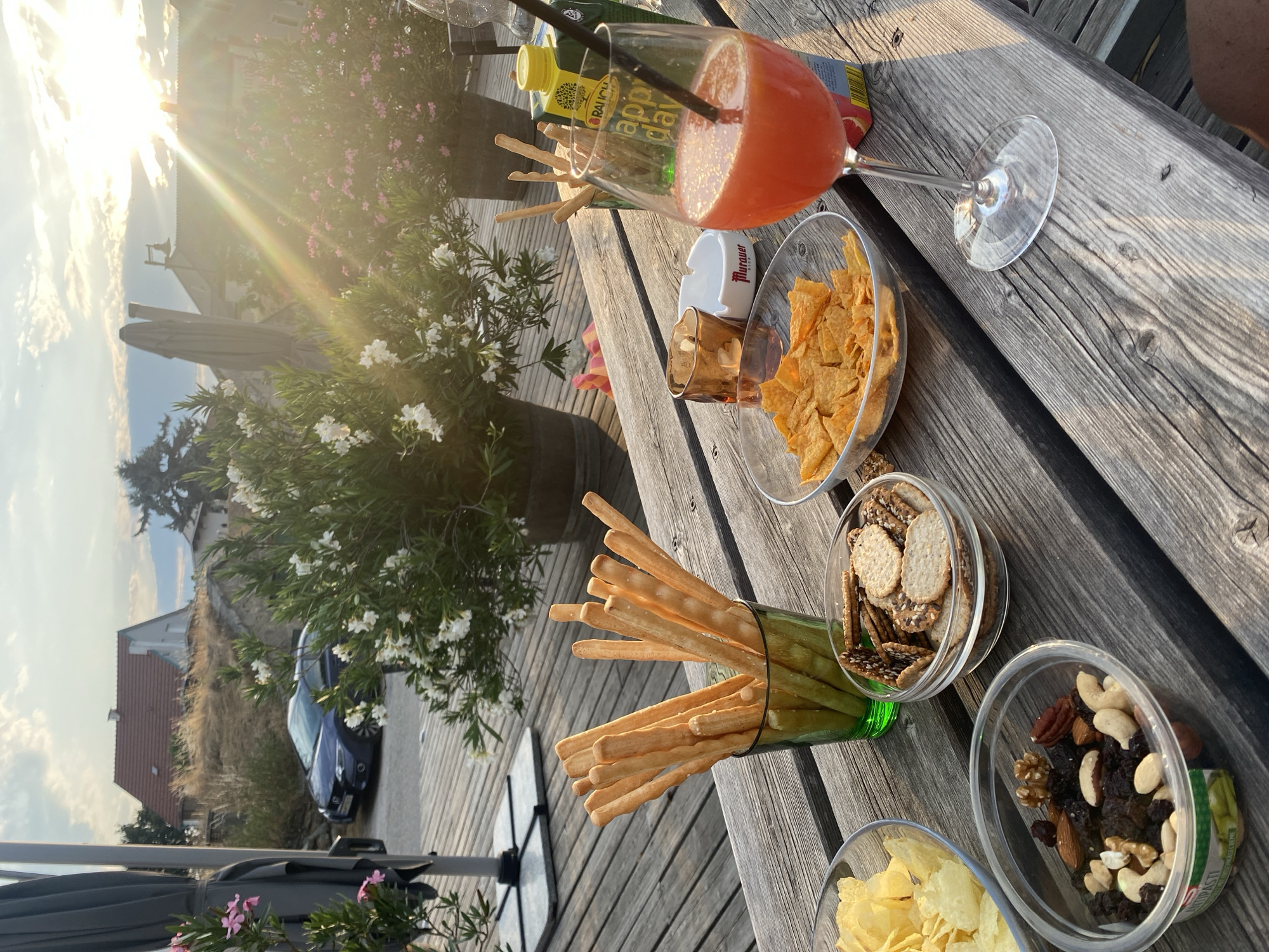 The image depicts an outdoor setting, likely a terrace or patio, with a rustic wooden table laden with snacks and a drink. In the foreground, several bowls contain a variety of snacks: potato chips, crackers, and a mix of nuts and dried fruits. A glass of red-orange liquid, presumably a cocktail or juice, is prominent, with a straw and a small ashtray nearby. Behind the snacks, a green glass holds a bunch of breadsticks. A carton of "Rauch Happy Day" juice is visible next to the drink. In the background, lush green plants with white and pink flowers are in bloom. Sunlight streams in from the right, creating lens flare and casting a warm glow. A blue car is parked in the distance, and a house with a red roof is situated on a gentle slope. The overall atmosphere suggests a relaxed, outdoor gathering. The presence of the "Murauer" brand on the ashtray suggests the location may be in or near Murau, Austria, although the prompt specified Purbach, Austria. Without further context or distinct landmarks, confirming the exact location within Purbach is not possible from the visual information alone.
