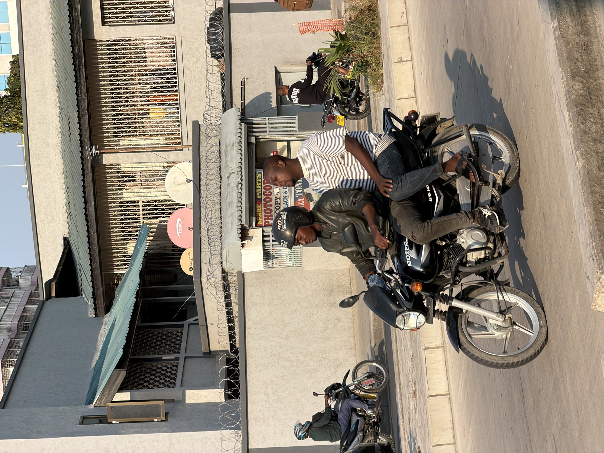 The image depicts a street scene in Dar es Salaam, Tanzania, with two men riding a motorcycle. The man in the front is wearing a black leather jacket and a black helmet, while the man behind him is wearing a striped t-shirt and blue jeans. The motorcycle is a black Boxer 125 HD. In the background, there is a building with a gray facade, a green corrugated metal roof, and barred windows. The building appears to be a commercial establishment with a sign that reads "SPARE PARTS PHOTOCOPY COPY & ... TONN". Another motorcycle is visible in the left background, with a rider wearing a helmet and a green jacket. Barbed wire is strung along the top of a wall in front of the building. The street is paved with asphalt and bordered by a concrete curb. The sun is low in the sky, casting long shadows.