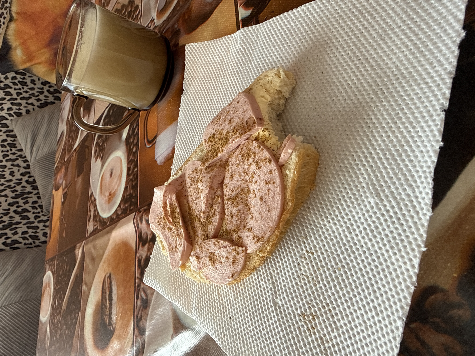 A breakfast scene is depicted indoors. A glass mug containing a light-colored beverage is placed on a tile surface patterned with coffee beans and espresso cups. To the right, a piece of bread topped with sausage slices, sprinkled with a fine powder, rests on a white paper napkin. The photograph also includes a section of bedding with a cheetah print and striped pattern. The location is unknown and the city of Sofia, Bulgaria, cannot be determined from the image.