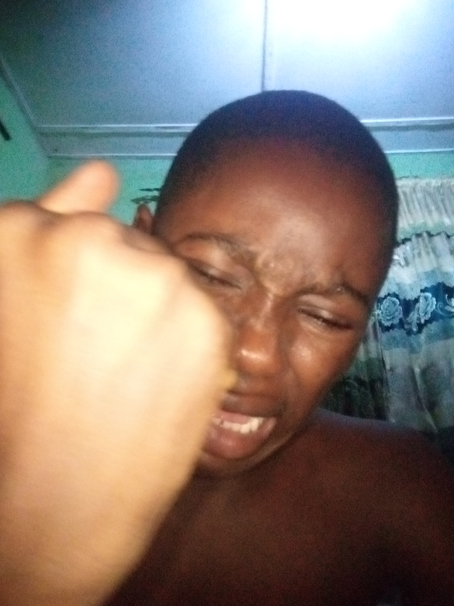 The image is a close-up, low-light selfie taken by a young person. The person's face is contorted in an expression that could be interpreted as pain or exertion, with their eyes squeezed shut and mouth open, revealing teeth. A hand is prominently in the foreground, obscuring a significant portion of the left side of the frame, and appearing to be raised towards the camera. The background is dimly lit, with what appears to be a curtain with a floral pattern visible on the right side. The overall scene is grainy and out of focus, suggesting it was taken with a low-resolution camera or in poor lighting conditions. There is no discernible information about the specific location within Shangisha, Nigeria, or any other contextual details of the environment.