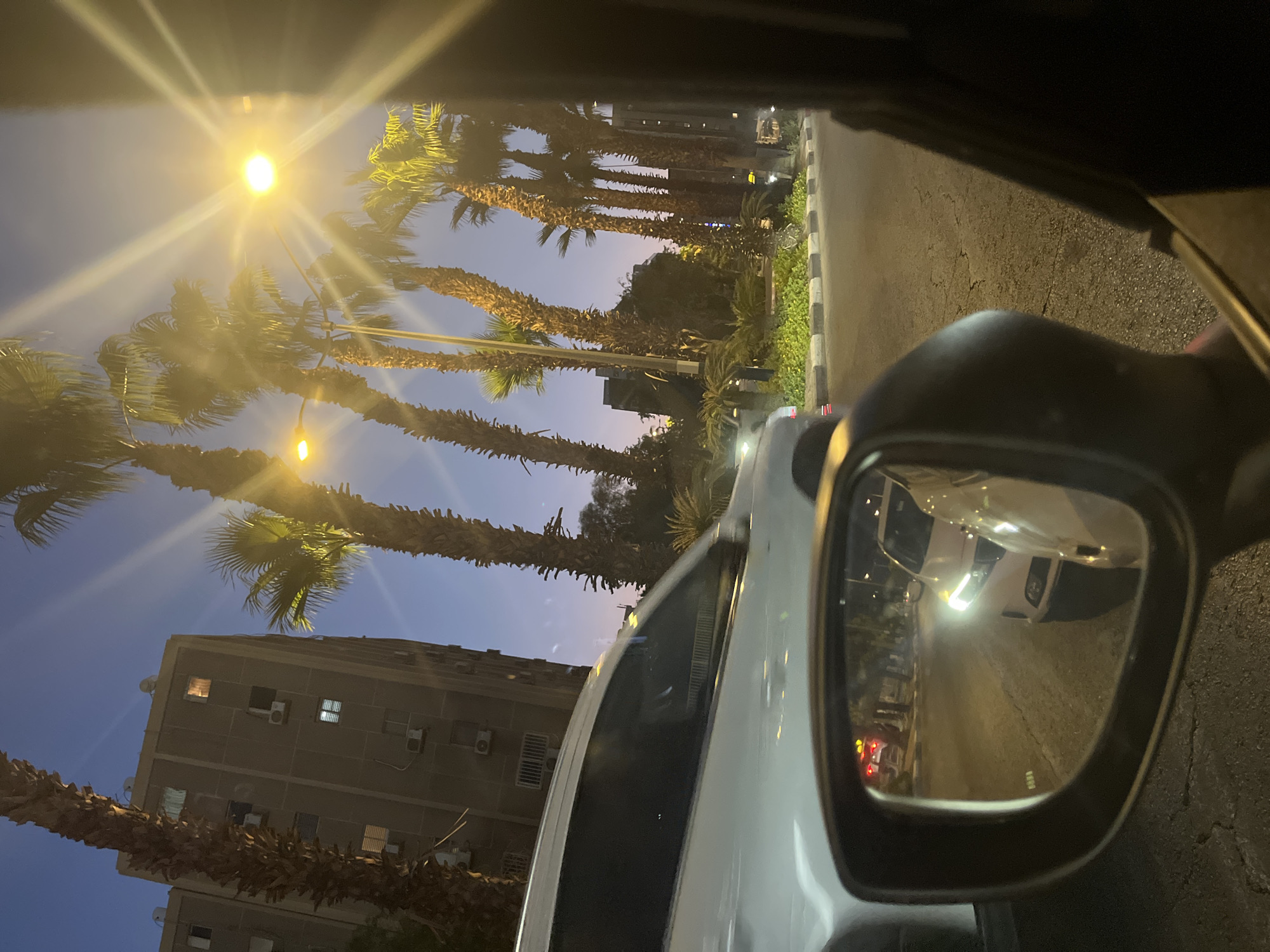 The image depicts a street scene at dusk or night, likely in a warm climate, given the presence of palm trees. A white car is the primary subject, seen from the perspective of someone inside another vehicle, looking out the side window and into the wing mirror. The car in front is partially visible, its sleek, modern design suggesting a contemporary model.
In the wing mirror, the reflection shows a white car with its headlights on, followed by another vehicle. The road surface appears to be asphalt, with some cracks visible. Along the side of the road, there is a raised median strip with green vegetation and small plants.
Behind the car, tall palm trees with their characteristic fronds dominate the skyline. Their rough trunks and sparse foliage are silhouetted against a deepening blue sky. In the background, a multi-story building with a somewhat utilitarian design is visible. It appears to be an apartment complex or office building, with several windows and air conditioning units mounted on its facade. Some windows are illuminated, suggesting occupancy.
Streetlights, emitting a bright yellow glow, illuminate the scene, casting long beams of light that create a starburst effect. The overall impression is that of a city street at night, with moderate traffic and ambient lighting. The image has a slightly grainy quality, indicative of low-light photography, and the colors are somewhat muted due to the time of day and artificial lighting.