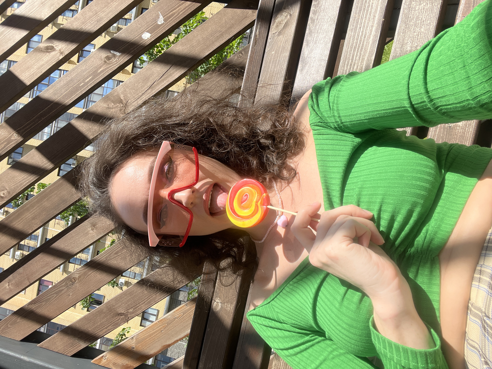 A young woman with dark, curly hair is sitting on a wooden bench, holding a lollipop to her mouth. She is wearing a bright green off-the-shoulder top and pink-tinted sunglasses. Behind her, a building with yellow brickwork and many windows is visible through a wooden slatted fence. The scene appears to be outdoors, possibly a residential courtyard in Kazan, Russia, given the architectural style of the building and the presence of outdoor furniture. The lighting suggests it is daytime.