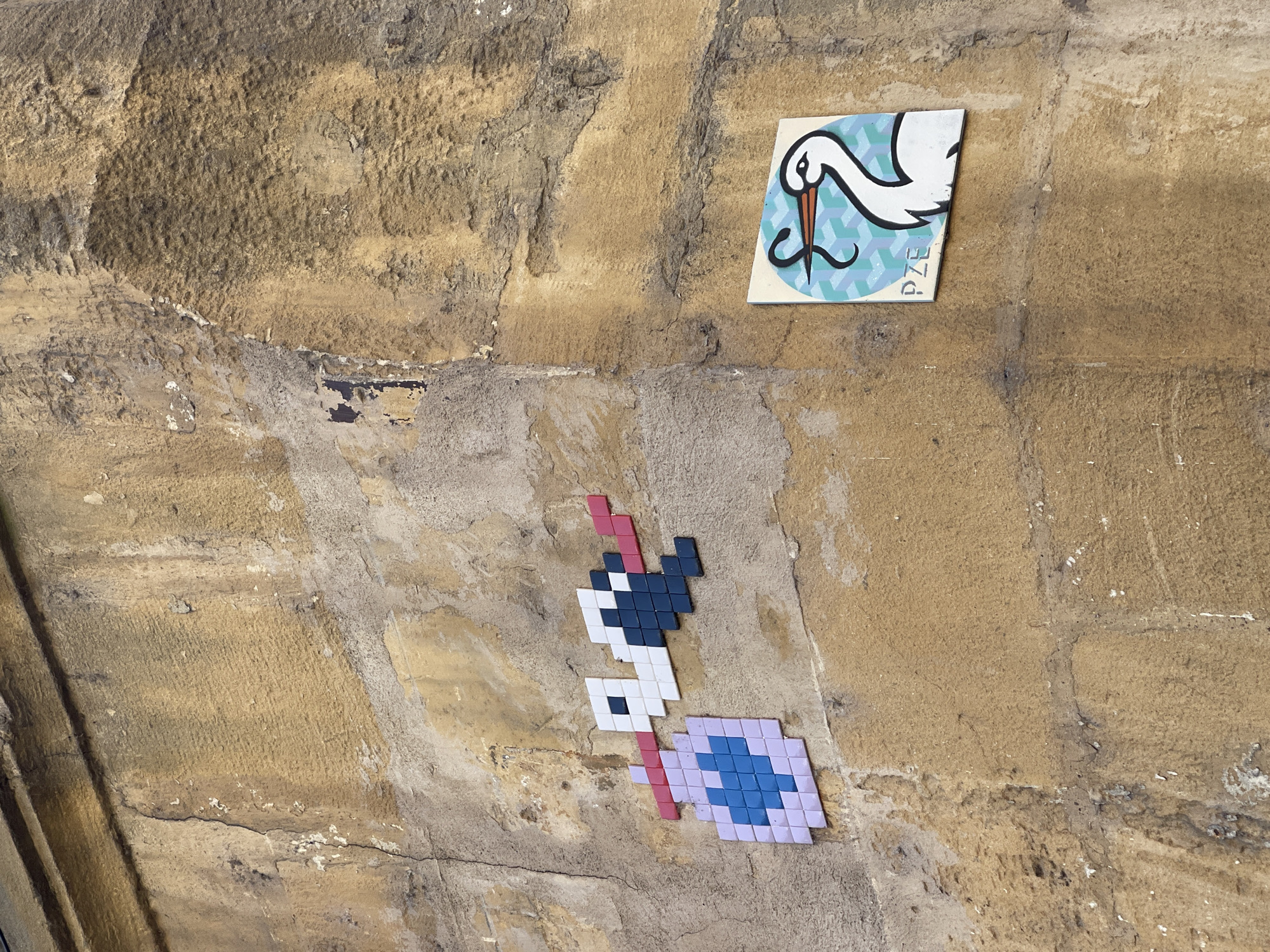 A close-up shows a tan, rough stone wall with two pieces of street art affixed to it. At the top right is a rectangular sticker depicting a white stork with a blue geometric background and the letters "PZE" printed on the lower right. Below the sticker is a piece of pixel art of a rabbit or other long-eared animal holding a blue and purple shield, constructed from small, colored tiles. This image was taken in the city of Metz, France.