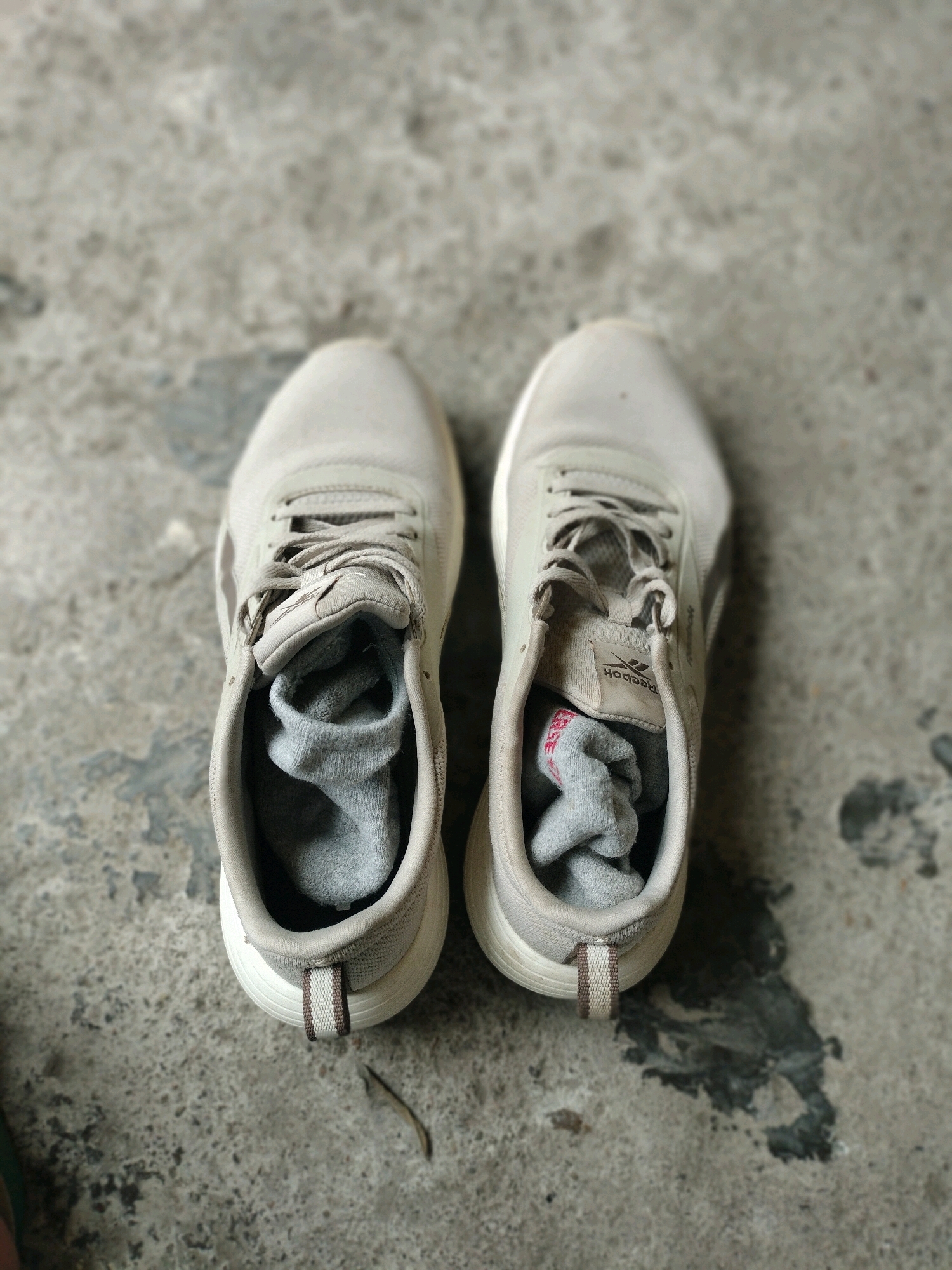 A pair of light gray athletic shoes with gray socks stuffed inside rests on a concrete surface. The shoes appear to be worn and are positioned side-by-side, with the left shoe slightly closer to the viewer. The concrete ground has a mottled texture with dark and light gray patches, suggesting it is an outdoor paved area. There is no indication of any specific location in Pembataan, Indonesia, within the image itself.