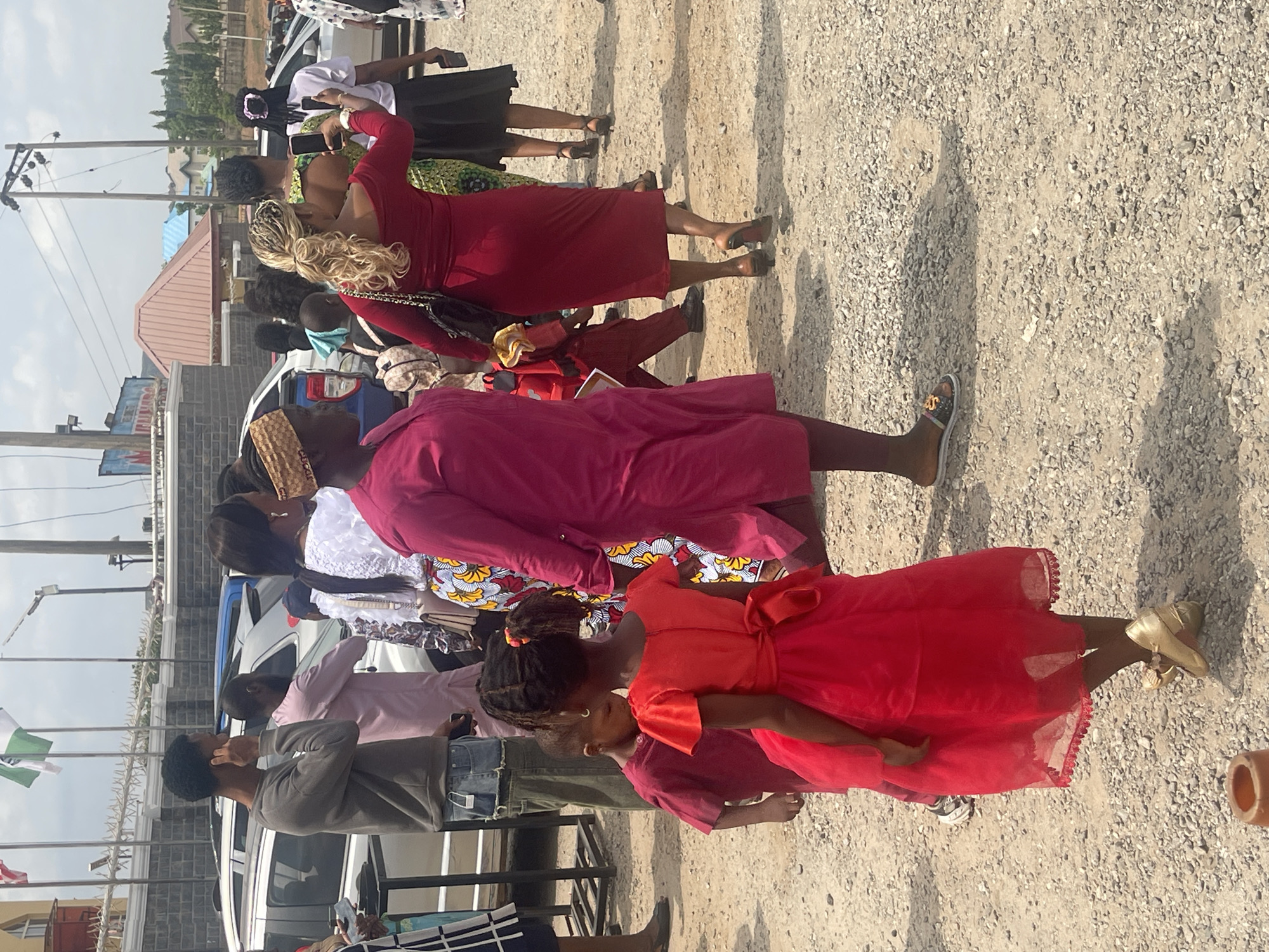 A group of people, including children in red dresses and adults in various clothing, are gathered on a gravelly surface outdoors. The scene appears to be in Abuja, Nigeria, based on the presence of Nigerian flags. Some people are standing, others are walking, and one woman is taking a photo with her phone. Cars are parked nearby, and buildings are visible in the background.