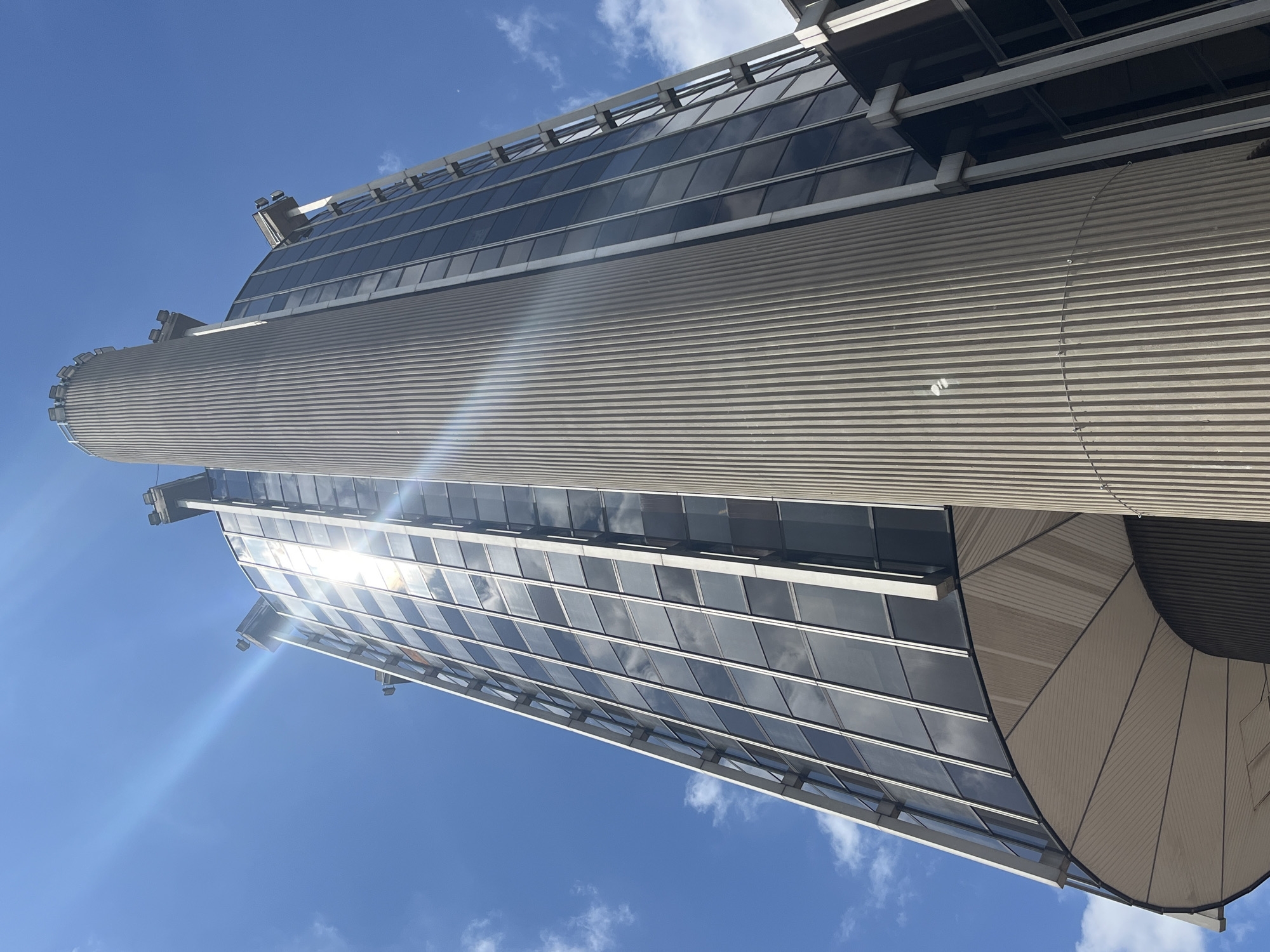 The image shows a low-angle, upward view of the Crédit Lyonnais building in Créteil, France. The building is a modern high-rise with a glass facade and a cylindrical concrete structure running vertically up its side. The sky is a clear blue with scattered white clouds. Sunlight glints off the building's windows, creating lens flare. The architectural style is characteristic of the late 20th century. There are no people visible in the image. The overall impression is one of urban architecture and a bright, sunny day.
