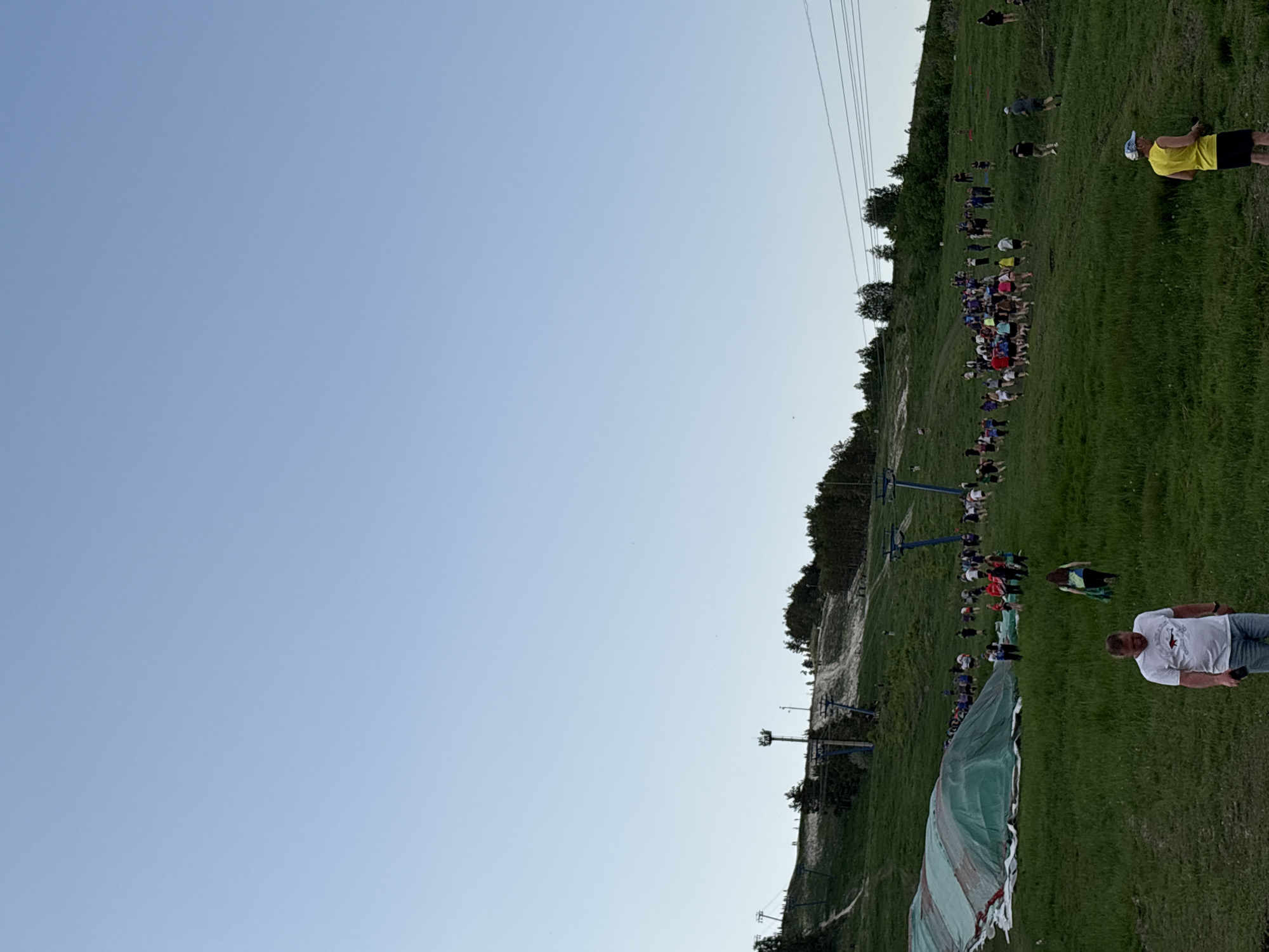 The image shows a grassy hillside in Khvalynsk, Russia. A large group of people is gathered on the slope. A ski lift is visible in the background. A large green and white tarp is partially visible in the lower right foreground. Individuals are standing and walking around. The sky is blue and clear. A man wearing a white shirt and jeans is standing in the foreground. Another person is wearing a yellow shirt and black shorts near the top right.