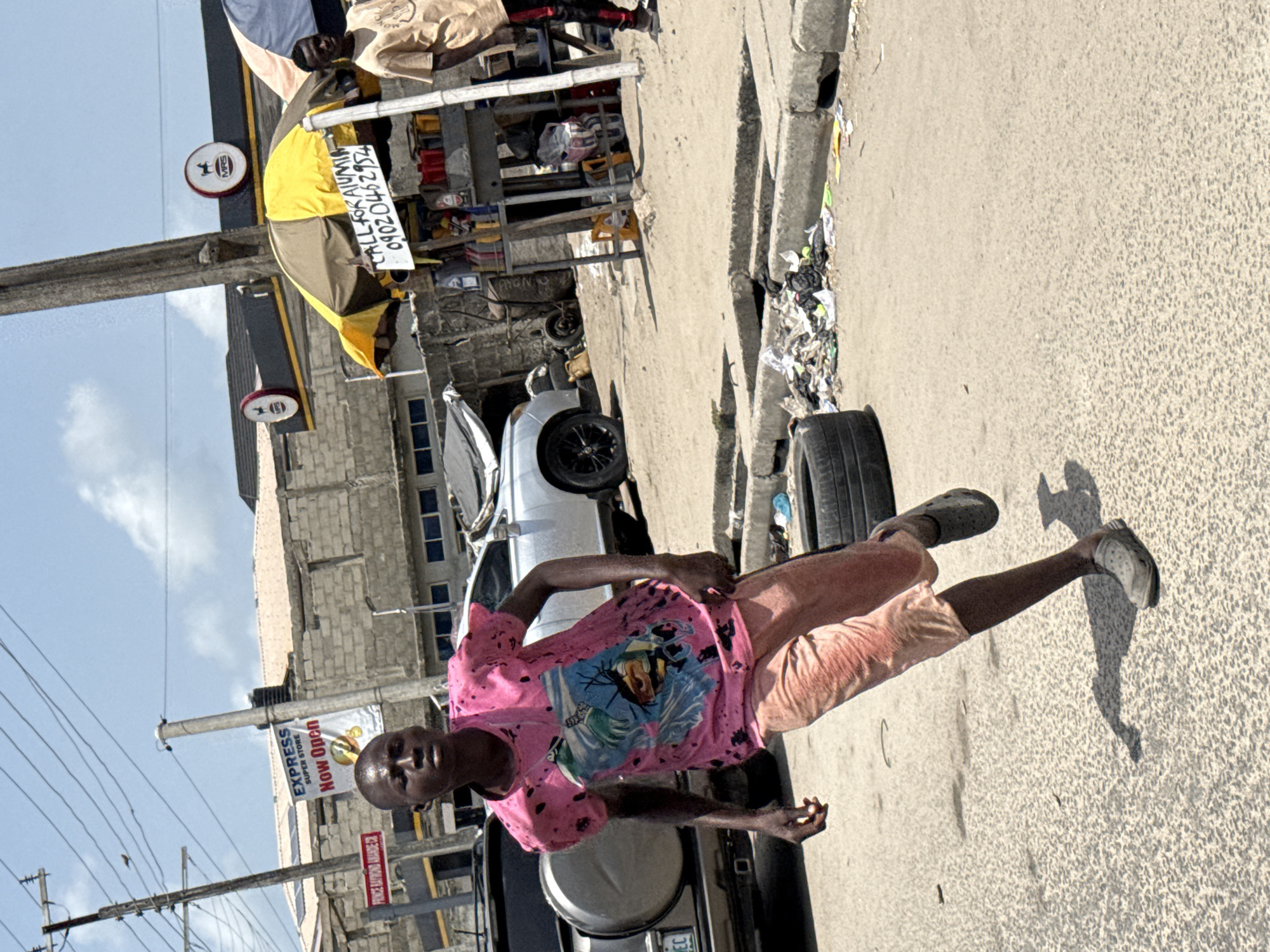 The image depicts a man in a pink t-shirt and salmon-colored shorts standing on a street in Ado, Nigeria. He wears dark footwear and casts a shadow on the paved road. Behind him is a vehicle, possibly a truck, with a visible license plate.
Further back, there's a silver-colored car and a makeshift market stall sheltered by a yellow umbrella with a sign advertising "CALL FOR ALUMIN 09020462954." Trash is visible along the curb.
Across the street, there is a building with a sign that reads "EXPRESS SUPER STORE Now Open," indicating a retail establishment. Another sign displays the street name "PRINCE RATROND ANANDE CR." Utility poles and power lines are visible against a bright sky. The scene appears to be a typical street setting in a commercial or residential area of Ado.