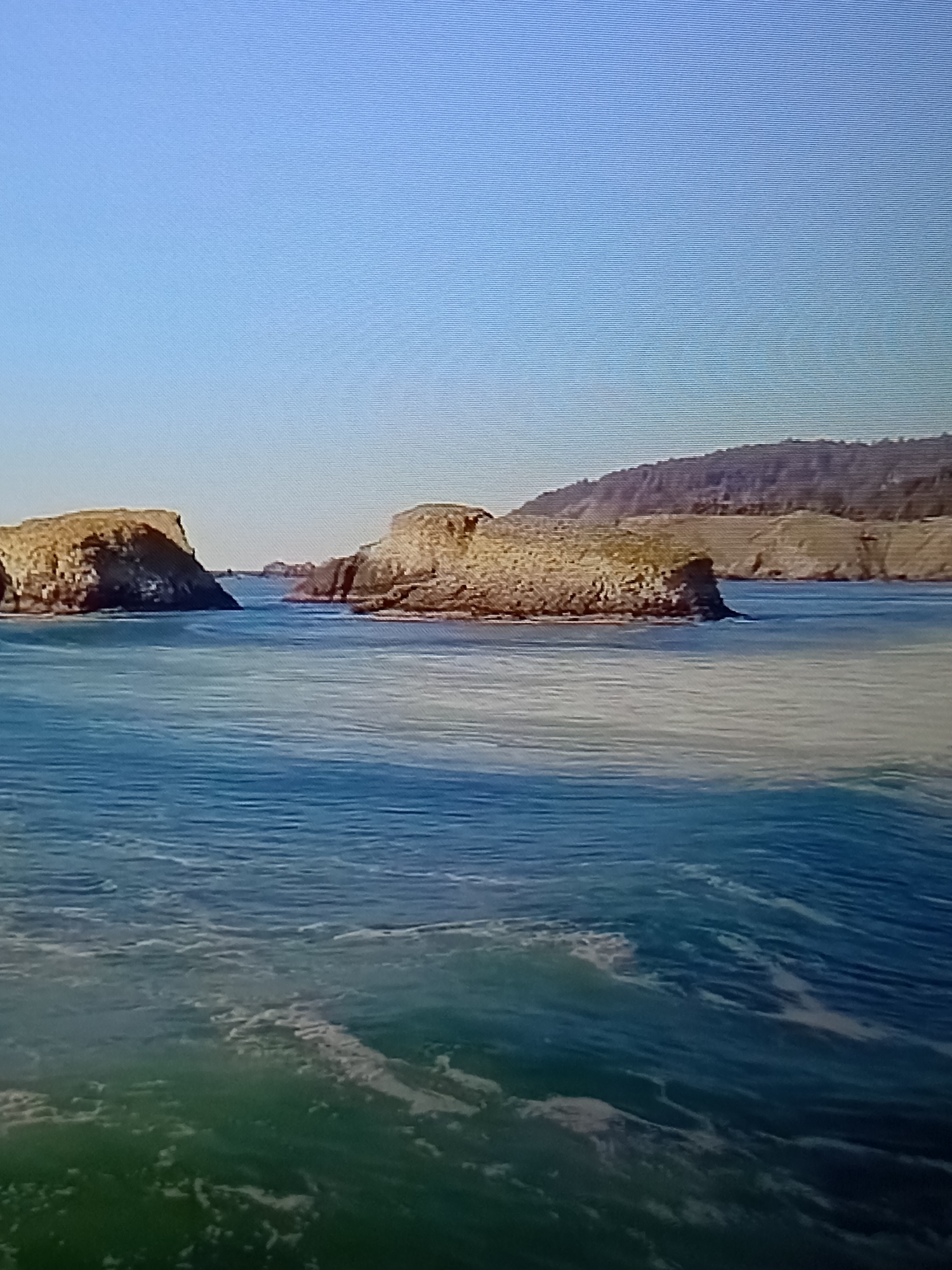 The image depicts a scenic natural landscape featuring rock formations rising out of a body of water under a clear blue sky. In the foreground, the water is a vibrant blue with white wave crests and darker green patches, suggesting depth and movement. Two prominent rock formations, light brown and layered in texture, emerge from the water. The larger one is on the left, with a steep, angular face. The other is to its right, appearing more rounded and broader. In the background, a rugged, brown-toned hillside or mountain range stretches across the horizon, partially obscured by haze. The overall impression is one of a serene, undeveloped natural environment. There is no indication of any urban setting or structures.