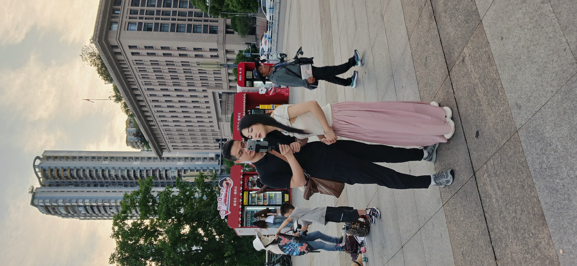 The image depicts a street scene in 江北城街道, China. A man and a woman stand together, the man holding a camera on a tripod, seemingly recording. The woman is dressed in a light-colored top and a long pink skirt. The man wears a black shirt and pants. Behind them is a row of small, red vending kiosks. A man with a camera, possibly a journalist, is nearby. Other pedestrians are present in the background. The environment includes tall buildings, trees, and paved ground. The sky is overcast.