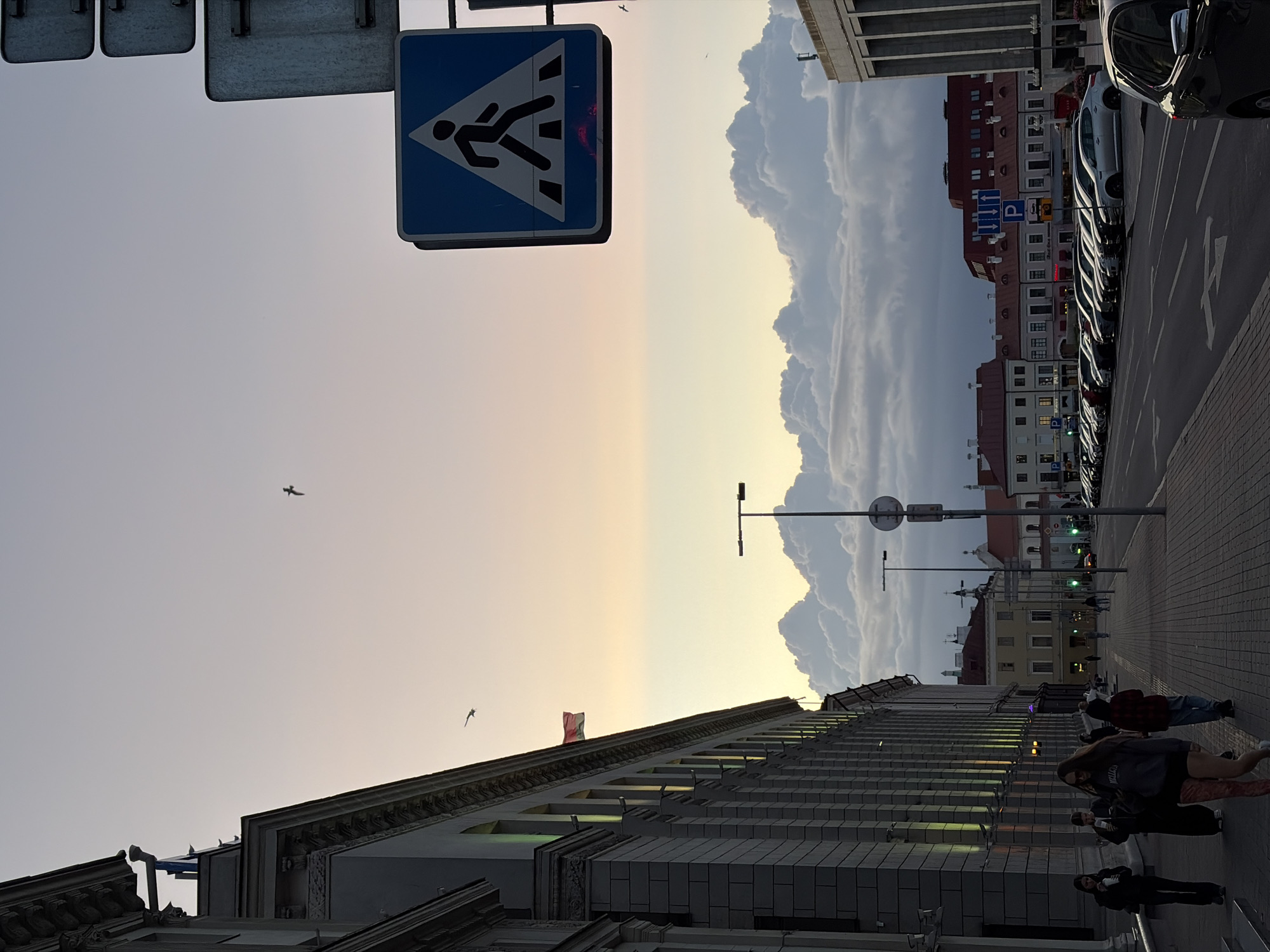 The image depicts a street scene in Minsk, Belarus, likely during the late afternoon or early evening. A large, grey, neoclassical building with arched windows and decorative columns dominates the left side of the frame. A Belarusian flag is visible on the building. On the right, a pedestrian crossing sign is prominently displayed, along with other street signs. A row of cars is parked along the street, with several buildings visible in the background, featuring red-tiled roofs. The sky above is a gradient of pale blue and soft orange, with dramatic, puffy white clouds. Several people are walking on the sidewalk, some engaged in conversation. Two birds are seen flying in the sky. The overall atmosphere is calm and urban.