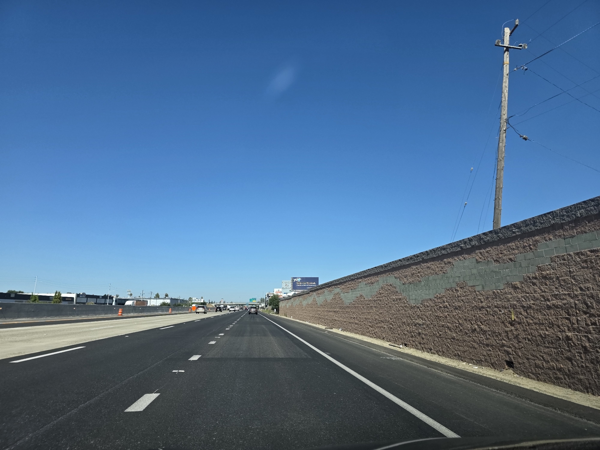 This is a first-person, forward-facing view from a vehicle traveling on a multi-lane highway in West Sacramento, California, United States of America. The highway has asphalt paving with white lane markings and a concrete barrier on the left. On the right, there is a textured brown retaining wall with a diagonal band of grey blocks. A wooden utility pole with wires stands against a clear blue sky. Distant buildings, some with advertisements on them, are visible on the left side of the highway. There are a few cars on the highway, all moving in the same direction as the viewer. The scene appears to be during daylight.