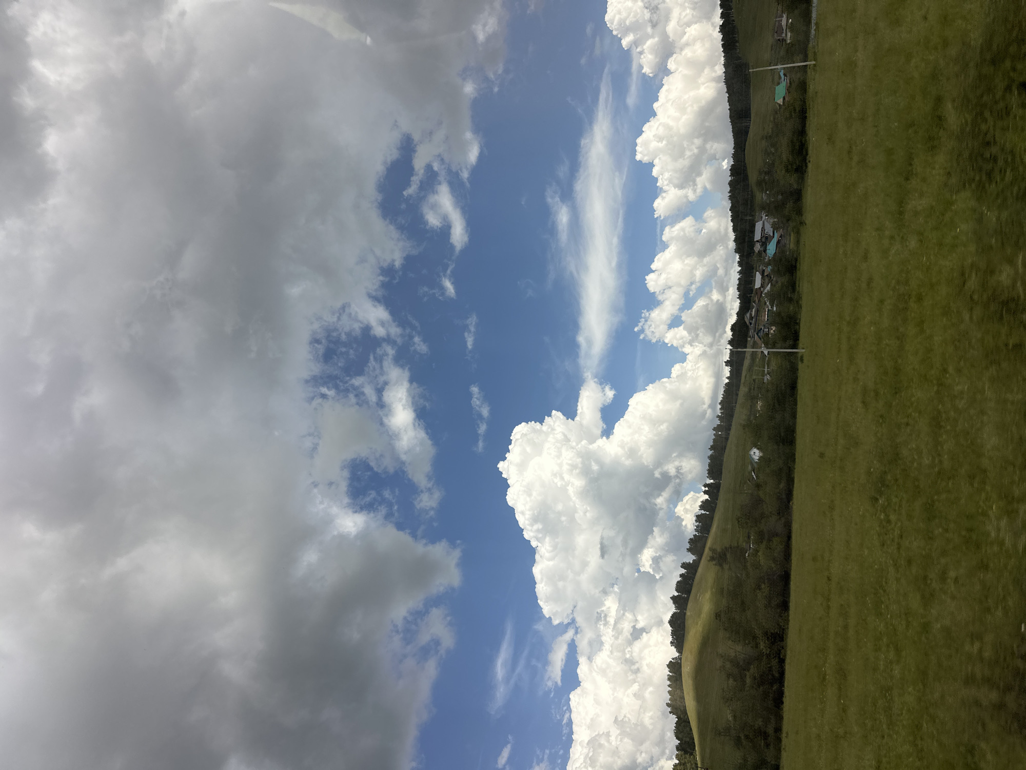 The image depicts a rural landscape under a dramatic sky. In the foreground, a vast expanse of green grass stretches towards a gentle hillside. The hillside is covered in trees and dotted with a small cluster of houses with brightly colored roofs, suggesting a village or settlement. A line of trees forms a dark band along the crest of the hill.
The sky is a striking feature, with a mix of bright blue sky and billowing white and grey clouds. The clouds are diverse in texture and shape, with some appearing fluffy and sunlit, while others are darker and more ominous. The overall impression is of a dynamic and possibly changing weather system. The image is taken from a slightly elevated perspective, possibly from a vehicle or higher ground. The scene is devoid of any people or animals.