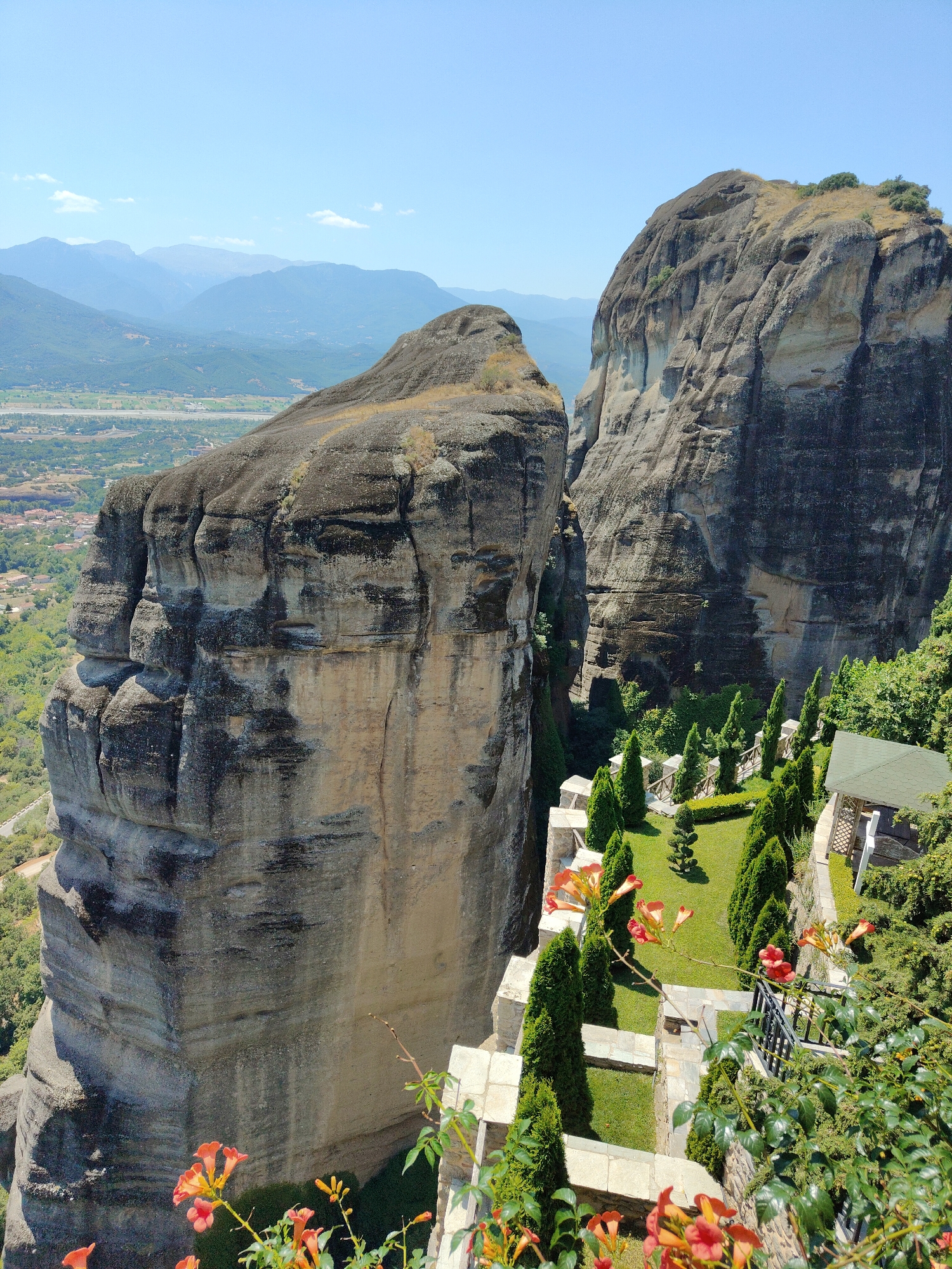 The image captures a scenic view of the Meteora rock formations in Kalambaka, Greece, under a clear blue sky. Towering, sheer rock cliffs dominate the foreground and midground, displaying textured surfaces with variations in light and shadow. In the midground and right side of the image, a well-maintained garden area is visible. This garden features neatly trimmed cypress trees, a manicured lawn, and a small gazebo with a green roof. A stone pathway and low stone walls delineate the garden spaces. Orange trumpet-shaped flowers bloom in the foreground and along the edge of the garden, adding a splash of color to the scene. The background reveals a hazy vista of rolling hills and distant mountains, with a glimpse of the town of Kalambaka nestled in the valley below. The scene is devoid of any people.