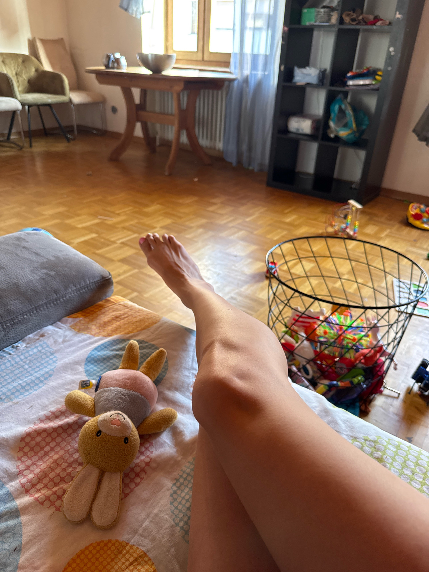 The image depicts an indoor scene, likely a living room or playroom, with a wooden floor and light-colored walls. In the foreground, a person's legs and right foot are visible, resting on a bed with a patterned duvet. A plush toy bunny is positioned on the duvet near the person's legs. To the right, a black wire basket overflows with colorful toys. Further in the background, a dark shelving unit is filled with various items, and a wooden table with chairs and a bowl are visible. A window with light blue curtains is also present. The image does not provide any information to confirm the location in Überherrn, Germany.