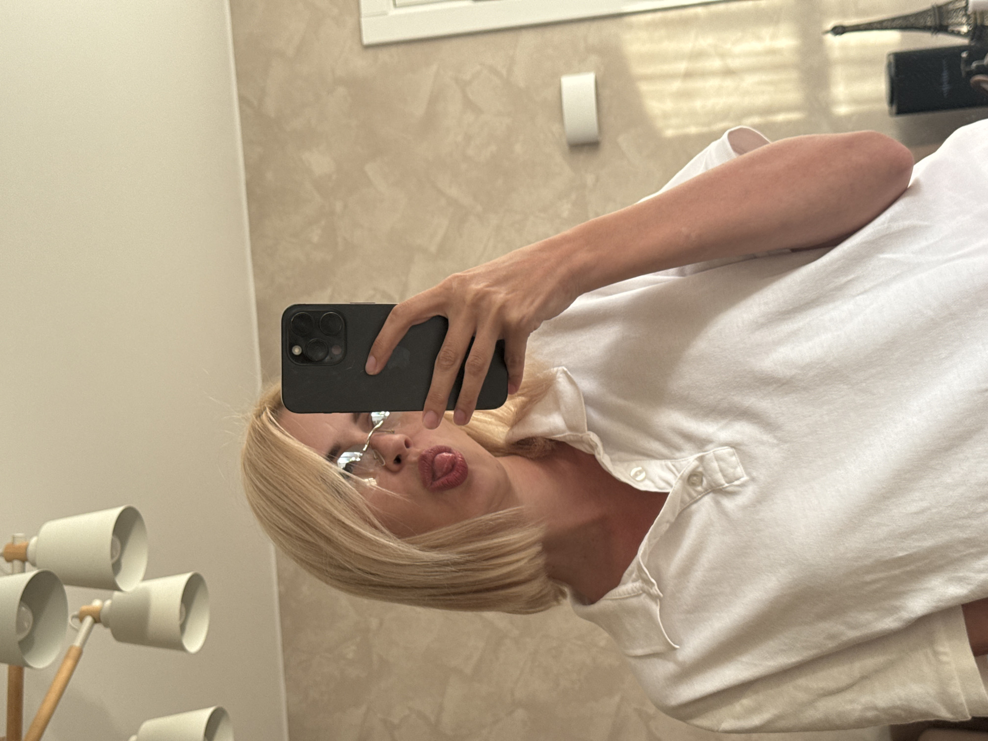 In an indoor setting, a person with short, blonde hair and glasses is taking a mirror selfie. They are wearing a white collared shirt and are sticking their tongue out at the camera. The background shows a textured wall and a modern light fixture. There is also a glimpse of a white rectangular object mounted on the wall and a small Eiffel Tower replica on a surface. The image appears to have been taken in an apartment in Saratov, Russia.
