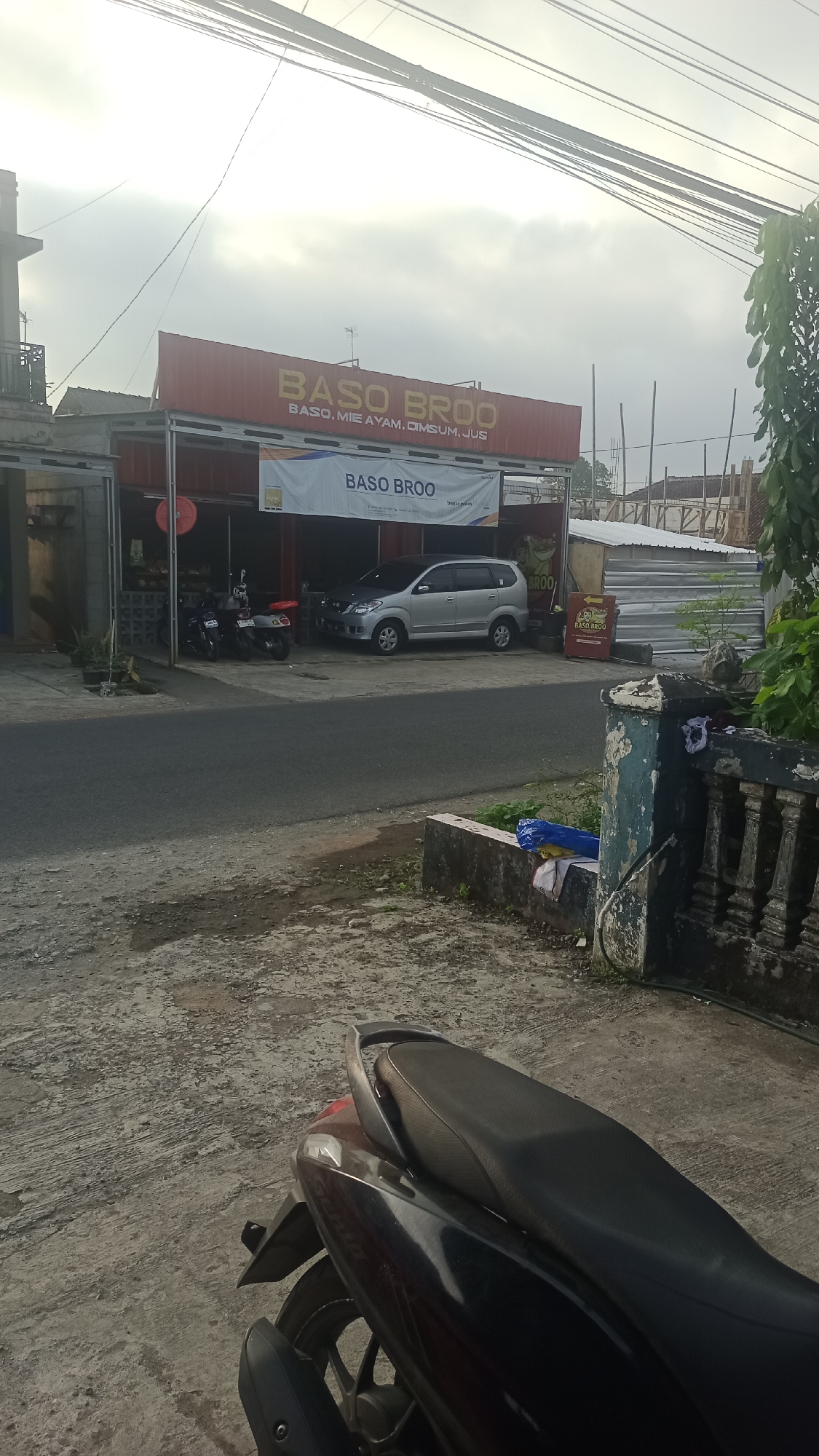 The image depicts a street view in Pakemitan, Indonesia. In the foreground, the rear section of a dark-colored motorcycle is visible, parked on a textured concrete surface. The street runs horizontally across the middle ground. On the opposite side of the street, a building with a red facade prominently displays a sign reading "BASO BROO" in yellow capital letters, with smaller text below indicating "BASO, MIE AYAM, DIMSUM, JUS". A banner featuring the "BASO BROO" logo is also hung on the building. A silver car is parked in front of the building, and two motorcycles are parked to the left of the building entrance. The environment appears to be an urban commercial area, with some construction or ongoing work visible further down the street. The sky is overcast, suggesting a cloudy day.