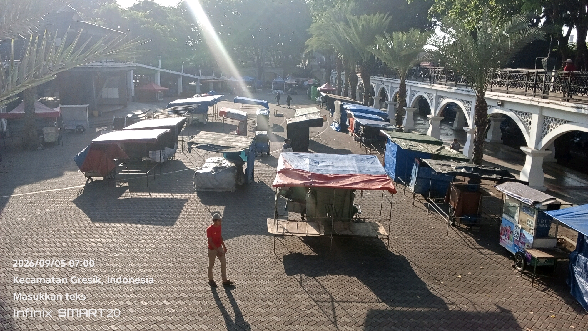 The image depicts an outdoor market scene in Gresik, Indonesia, on September 5th, 2026, at 7:00 AM. The market appears to be in a state of preparation or early operation, with numerous vendor stalls set up. Many of the stalls are covered with tarpaulins in various colors, including blue, red, and grey. Some stalls are empty, while others appear to be covered with protective materials, suggesting they may be closed or awaiting goods.
A single individual, a man wearing a red shirt and tan pants, is walking across the paved area of the market. He is casting a long shadow, indicating the sun is low in the sky. In the background, there is a white arched structure with decorative railings, possibly a bridge or elevated walkway, with palm trees lining it. Other tents and structures are visible in the distance, suggesting a larger market area. The ground is paved with rectangular interlocking bricks. Text overlays on the image indicate "2026/09/05 07:00", "Kecamatan Gresik, Indonesia", "Masukkan teks", and "Infinix SMART 20".