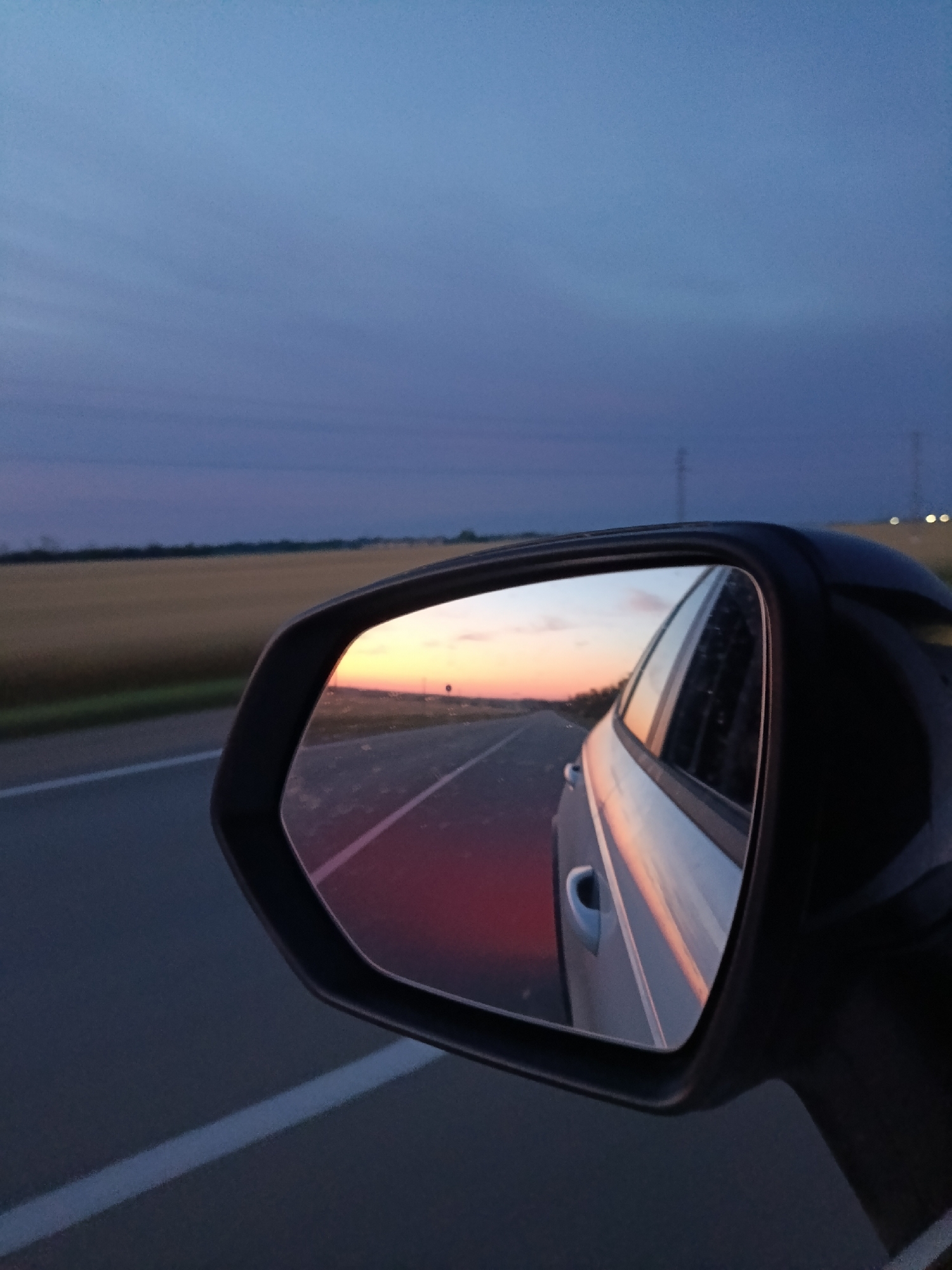 The image depicts a rear-view mirror reflection of a road at dusk. The sky transitions from a deep blue to lighter shades of purple and orange, indicating the sunset. A car is visible in the reflection, as is a road with white lines. Fields of dry grass or crops are on either side of the road. Electrical poles are visible in the distance. The photograph appears to be taken from within a vehicle, likely in a rural or suburban area. No specific landmarks in Staroderevyankovskaya, Russia are identifiable.
