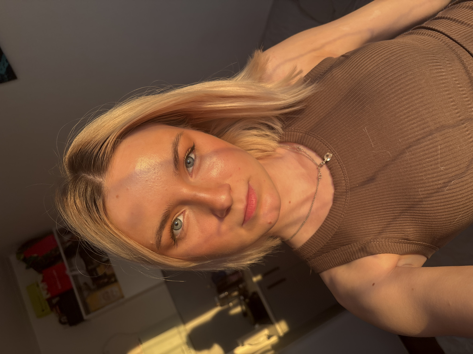 A candid portrait features a young woman taking a selfie in an indoor setting, illuminated by warm, golden light suggestive of late afternoon or early morning sun. The image is angled, implying the phone was held upright while the subject was reclining or tilting her head.
The primary subject is a woman with light skin, short to medium-length blonde hair, and striking blue-green eyes. She has a neutral expression, with subtle makeup highlighting her lips and eyes. She is wearing a simple brown ribbed tank top or camisole, and a delicate silver necklace with a small clear pendant. Several fine, light-colored hair strands are visible on her top.
The lighting is a prominent feature, creating strong highlights on her forehead, nose, and cheekbones, as well as catching the light in her blonde hair. The background is softly blurred but reveals hints of an indoor environment, including dark furniture or walls and what appear to be shelves with items like books or folders. No specific activity beyond taking the selfie is depicted, suggesting a casual, personal moment. There is no visible text in the image.