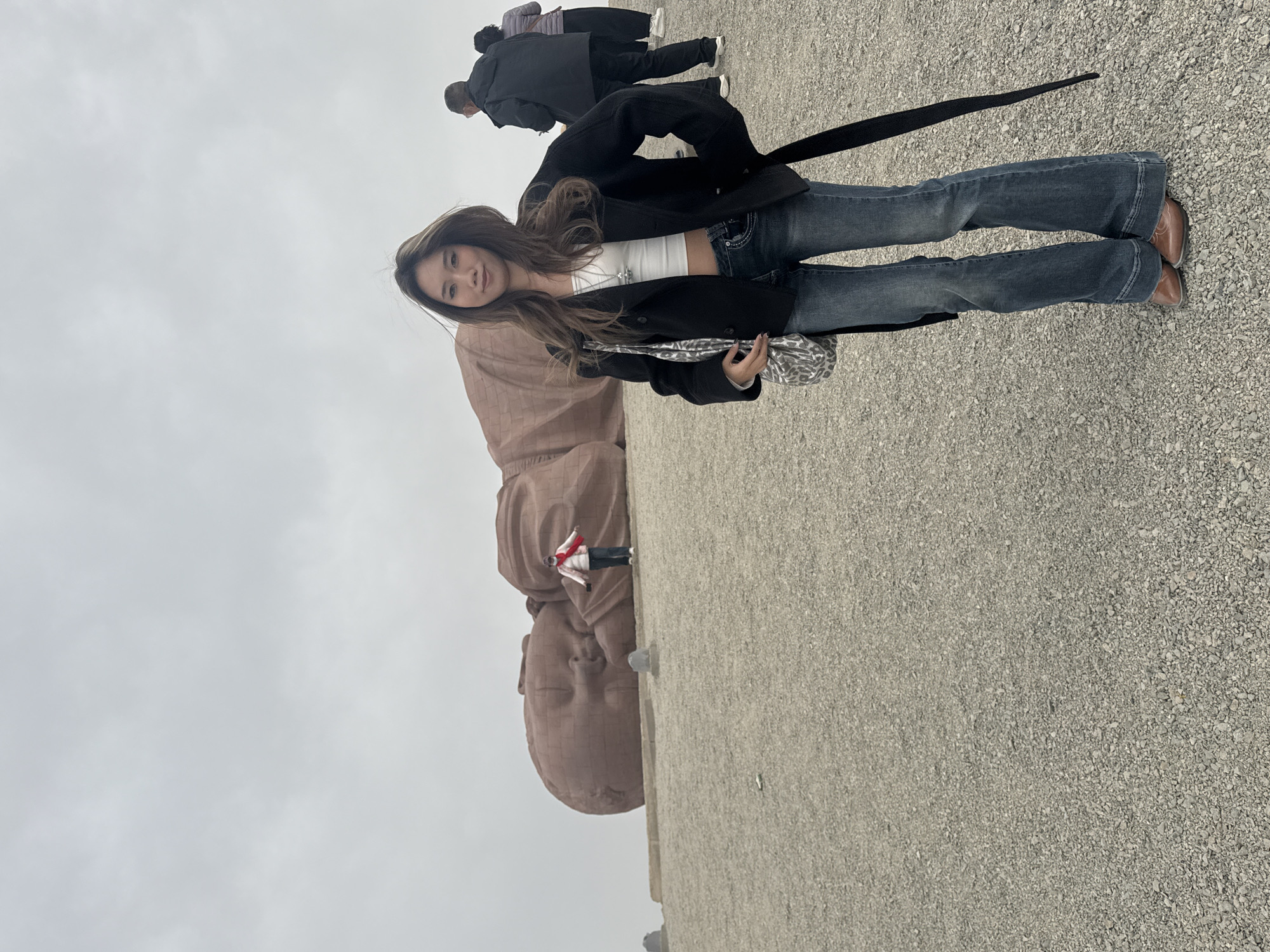 The image shows a woman standing in front of a large, tan-colored sculpture of a baby's head in Guazhou County, China. She has long brown hair, wears a white crop top, blue jeans, and a black jacket. She is holding a patterned bag and smiling. In the background, there are a few people walking. The ground is covered in small rocks and the sky is overcast.