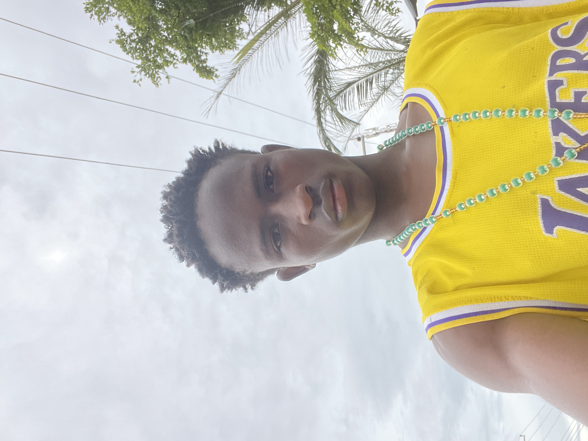 The image shows a young Black male from the chest up, looking directly at the camera. He has short, dark, curly hair and is wearing a bright yellow basketball jersey with purple and white trim. He is also wearing a long, beaded necklace in shades of green and gold. The background is overcast with a few power lines visible, and a palm tree and other greenery can be seen to the right. The lighting suggests it is daytime. The man's expression is neutral. The image appears to be a selfie. The specific location in Jos, Nigeria, is not identifiable from the visual information provided.