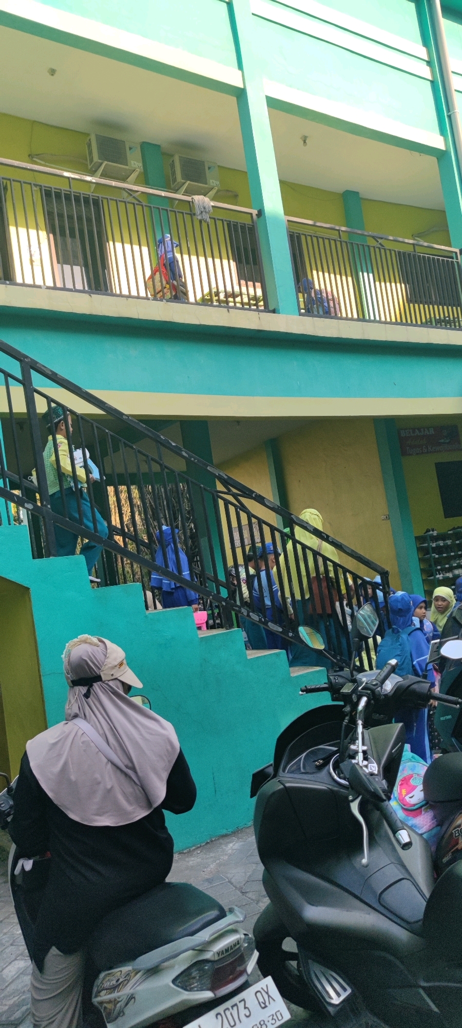 In Banjarsari, Indonesia, an outdoor scene depicts a bright, multi-story building with turquoise and yellow accents. A group of children, dressed in school uniforms and headscarves, are descending a set of outdoor stairs, some carrying bags. A person wearing a headscarf and covering their face is seated on a motorcycle in the foreground, facing away from the camera. The building appears to have balconies with metal railings, and air conditioning units are visible on an upper level. The overall impression is of a school environment during what seems to be dismissal or arrival.