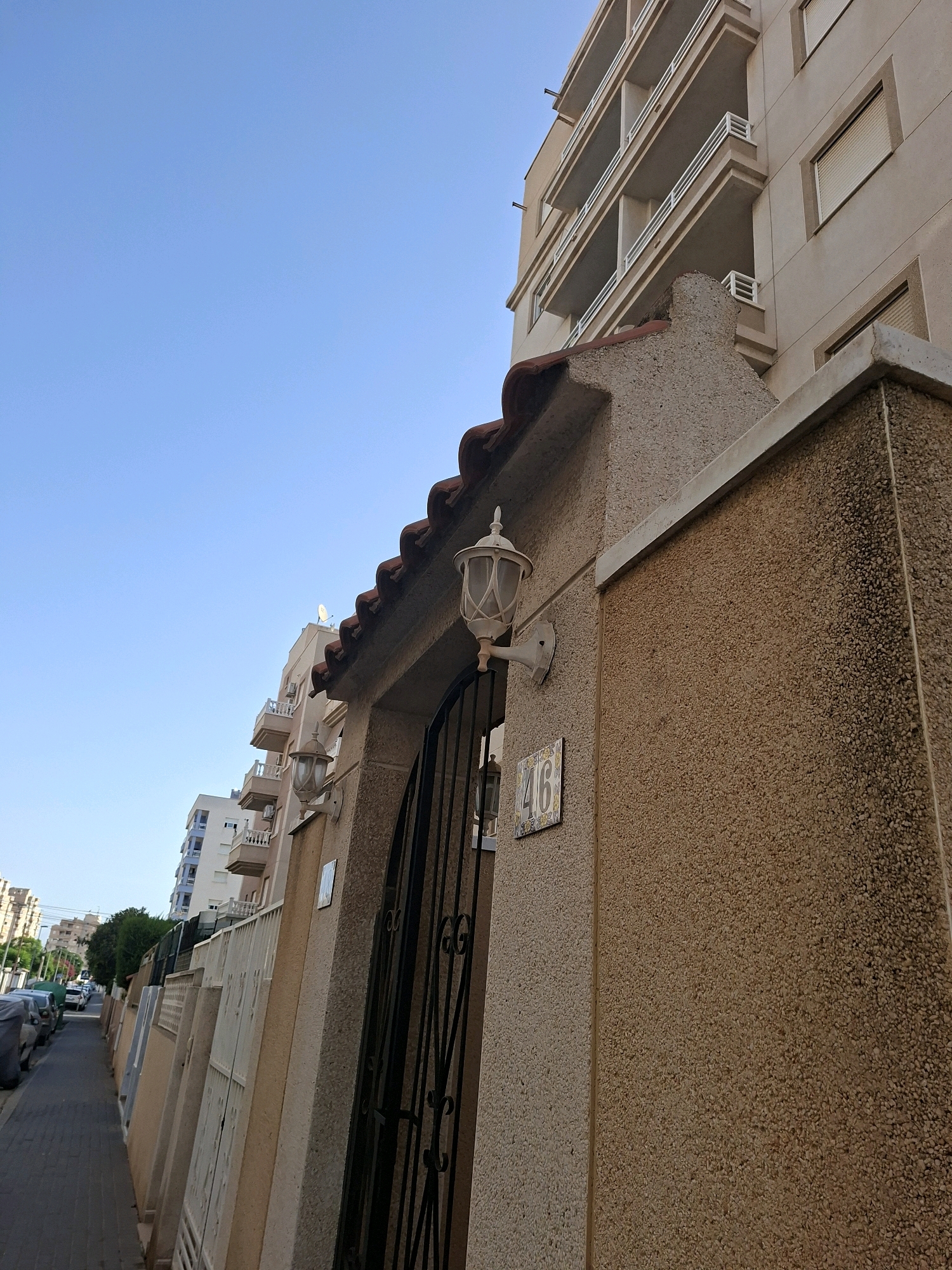 The image captures a street scene in Torrevieja, Spain, with a focus on a building facade and its entrance. A clear blue sky dominates the upper portion of the frame. In the foreground, a textured, light brown stucco wall is visible, leading to an arched entryway with a black wrought-iron gate. A white lantern is mounted on the wall next to the entrance, and a tile with the number "46" is affixed below it. The roofline above the entrance features reddish-brown tiles.
To the left of the main building, a sidewalk lined with parked cars runs along the street. Further down the street, a multi-story building with white balconies and beige siding is visible. Additional apartment buildings and some greenery are present in the background. The overall scene depicts a residential street on a sunny day in Torrevieja.