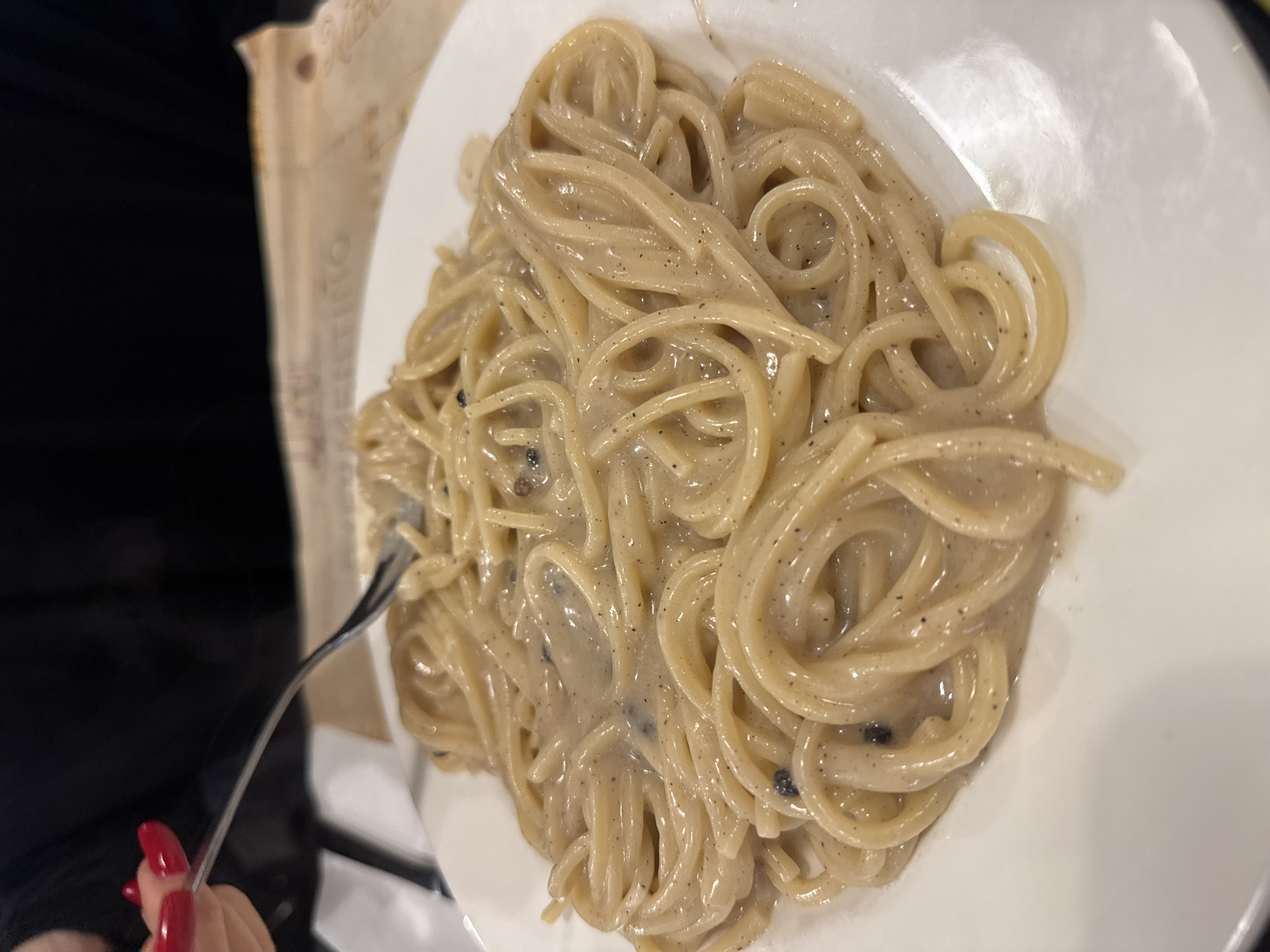 A close-up, slightly elevated shot captures a white, round plate filled with spaghetti. The pasta is generously coated in a viscous, light beige sauce speckled with fine black pepper, characteristic of a Cacio e Pepe dish. A silver-colored fork is positioned within the spaghetti, held by a left hand visible in the upper left of the frame. The hand displays manicured fingernails painted a bright red. The individual holding the fork is wearing a dark, possibly black, long-sleeved garment. The background is softly blurred, revealing a light brown paper object, possibly a placemat or menu, with partially legible text including "ZETTINO." The setting is an indoor dining environment, likely a restaurant table. Based on the traditional Roman specialty being consumed, the location is the city of Rome, Italy.