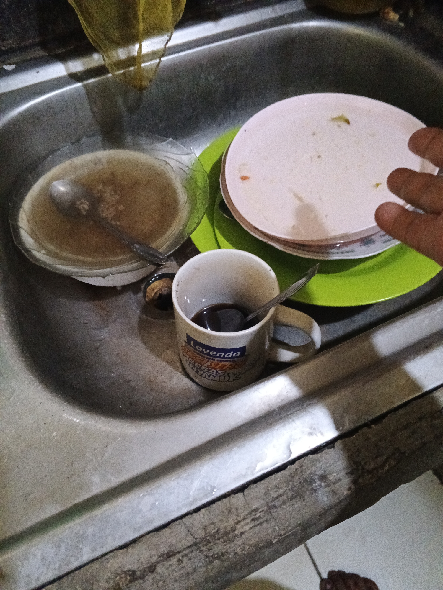 In a kitchen sink in Warungkiara, Indonesia, dirty dishes are piled up. A glass bowl contains a spoon submerged in murky liquid, possibly soapy water, with small food particles visible. Next to it, a stack of two white plates sits on a green plastic plate. The top plate shows remnants of food. A white ceramic mug, with the word "Lavenda" visible on its side, is filled with dark liquid, likely coffee, and a spoon rests inside. A hand, with fingers slightly curled, enters the frame from the right, appearing to reach for the dishes. The sink itself is made of stainless steel and shows signs of recent use, with water droplets and residue. The surrounding environment appears to be a domestic kitchen, with a wooden surface visible below the sink.