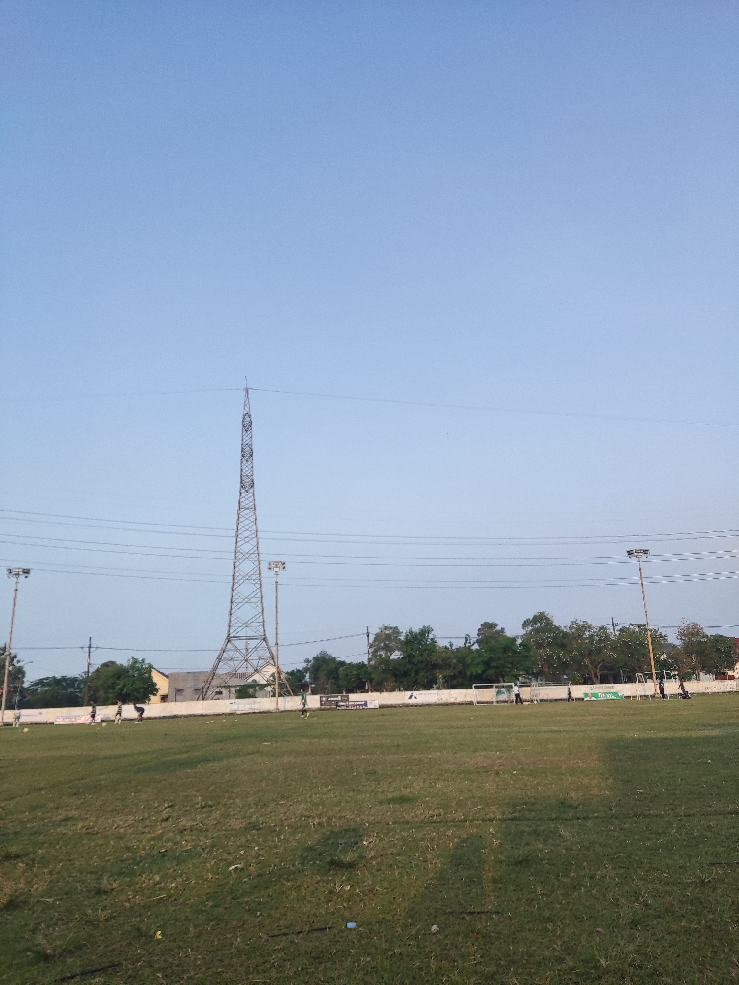 The image depicts an outdoor soccer field in Sidokepung, Indonesia. The field is largely green with some patches of dry grass. In the background, there is a white wall with a fence behind it. Several trees are visible beyond the fence, along with buildings and a tall, latticed transmission tower. Two floodlights are also present, one on the left and one on the right side of the frame. Several individuals, appearing to be young people, are scattered across the field, some engaged in what looks like a soccer practice or game. There are soccer goals at either end of the field. The sky is clear and pale blue, indicating daytime. The overall impression is a typical community sports ground.
