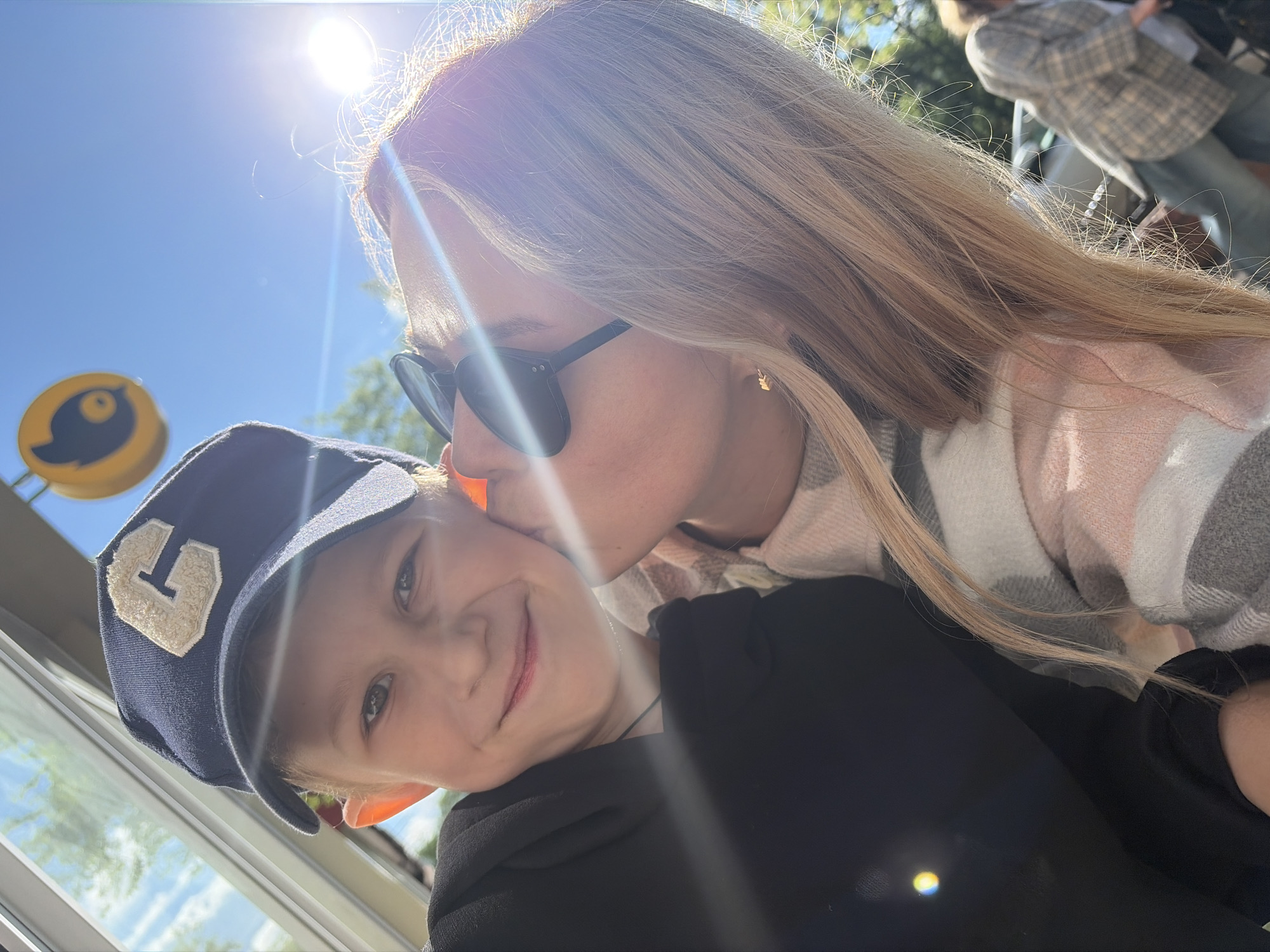 A woman wearing sunglasses and a pink and gray plaid scarf is kissing a young boy on the cheek. The boy is wearing a navy blue baseball cap with a white "G" on the front. They appear to be near a window. Outside the window is a yellow sign with a bird symbol, a Metro sign and a person walking by. Based on the Metro sign, the location is likely Chaikovsky, Russia.