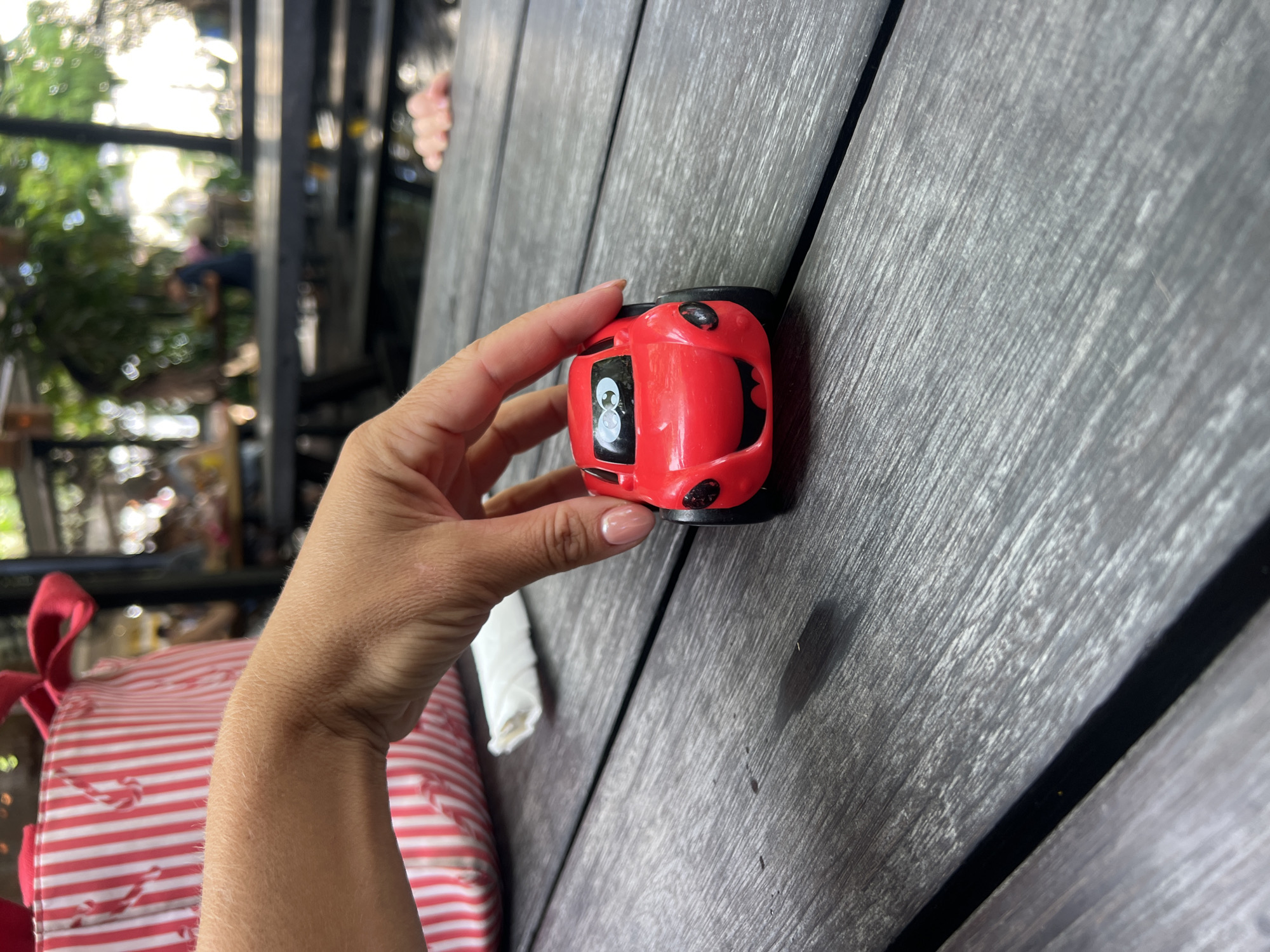 A person's hand is holding a red toy car on a wooden table. The car has a cartoonish face with large eyes and a smiling mouth. In the background, there are blurred images of people sitting at tables in what appears to be an outdoor dining area. The environment suggests a casual setting, possibly a cafe or restaurant, in North Kuta, Indonesia.