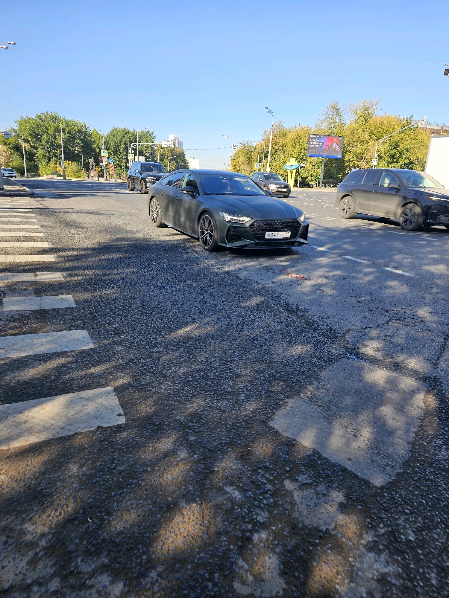 The image captures a street scene in Moscow, Russia, under a clear blue sky. A dark green Audi sedan is positioned in the foreground, facing right, stopped on a crosswalk. Its license plate reads "B841TY 99". To the right of the Audi, a black Audi station wagon is also stopped on the road. Further down the street, other vehicles and pedestrians are visible. Trees line the sides of the road, and a billboard advertising a bank with a 1% offer is prominent in the background. The road surface appears wet in places, and shadows from trees are cast across the asphalt. A small patch of reddish-pink material, possibly blood, is visible on the road in front of the dark green Audi, suggesting a recent incident.