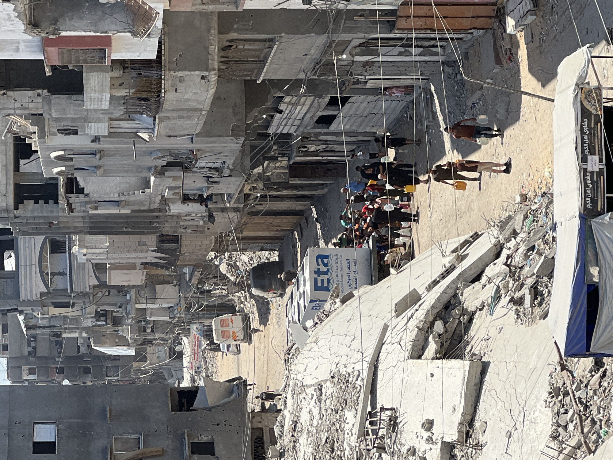 The image depicts a street scene in Gaza City, Palestinian Territories, characterized by widespread destruction of buildings. In the foreground, a large pile of rubble and concrete debris dominates the left side of the frame, with a partially visible blue and white tarp-covered structure in the bottom left corner. A white water tank with "Eta" branding and Arabic text is visible on a truck amidst the debris.
The street itself is dusty and littered with debris. A number of people are walking along the street and gathered in small groups. Some individuals are carrying containers, suggesting the transport of water or other essential supplies. The buildings lining the street show significant damage, with collapsed sections, exposed rebar, and missing walls. The architecture appears to be multi-story residential or commercial buildings. Overhead power lines are strung across the street, some of which appear damaged or dangling.
In the background, vehicles, including a white van and a dark-colored van, are present on the street. The overall atmosphere suggests a community coping with the aftermath of conflict or disaster, with a focus on survival and essential resource management. The sunlight indicates it is daytime.