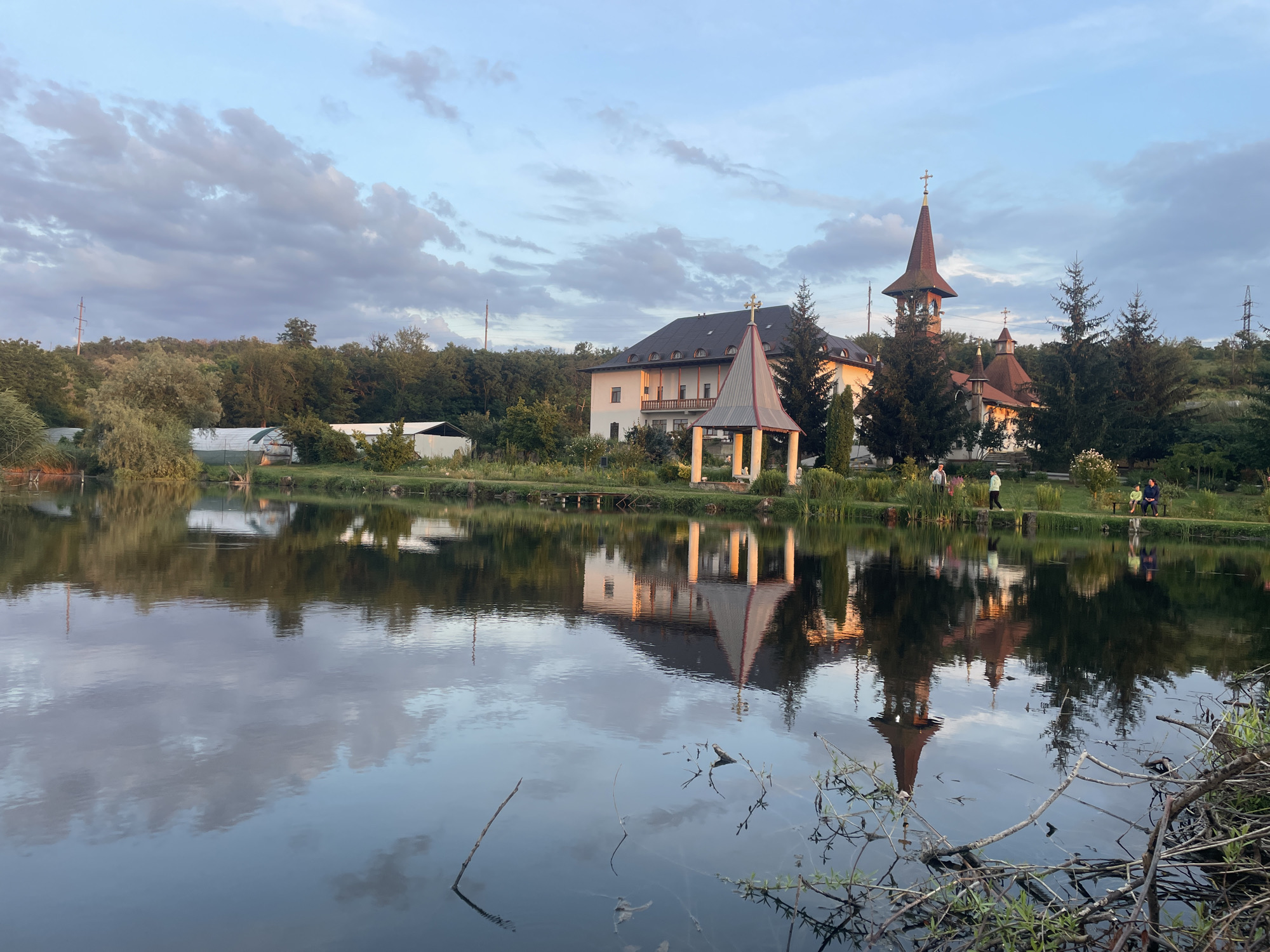 The image depicts a tranquil scene featuring a serene lake reflecting the sky and surrounding landscape. On the far bank of the lake, a complex of buildings, including a church with a distinctive spire and a larger, multi-story structure, are visible. These buildings appear to be part of a monastery or religious complex, with architectural elements suggesting Eastern European or Orthodox Christian influences. The sky above is a mix of blue with scattered clouds, hinting at either early morning or late afternoon light. Lush greenery, including trees and bushes, lines the bank of the lake and extends up a gentle incline behind the buildings. Several figures are scattered along the bank, some walking and others seated, suggesting a public or recreational area. The water itself is calm, creating a mirror-like reflection of the structures, trees, and clouds. The foreground of the image shows the edge of the lake with some dry branches and vegetation. Without specific landmarks visible in the image, it is not possible to definitively identify the location within the city of Cahul, Moldova.