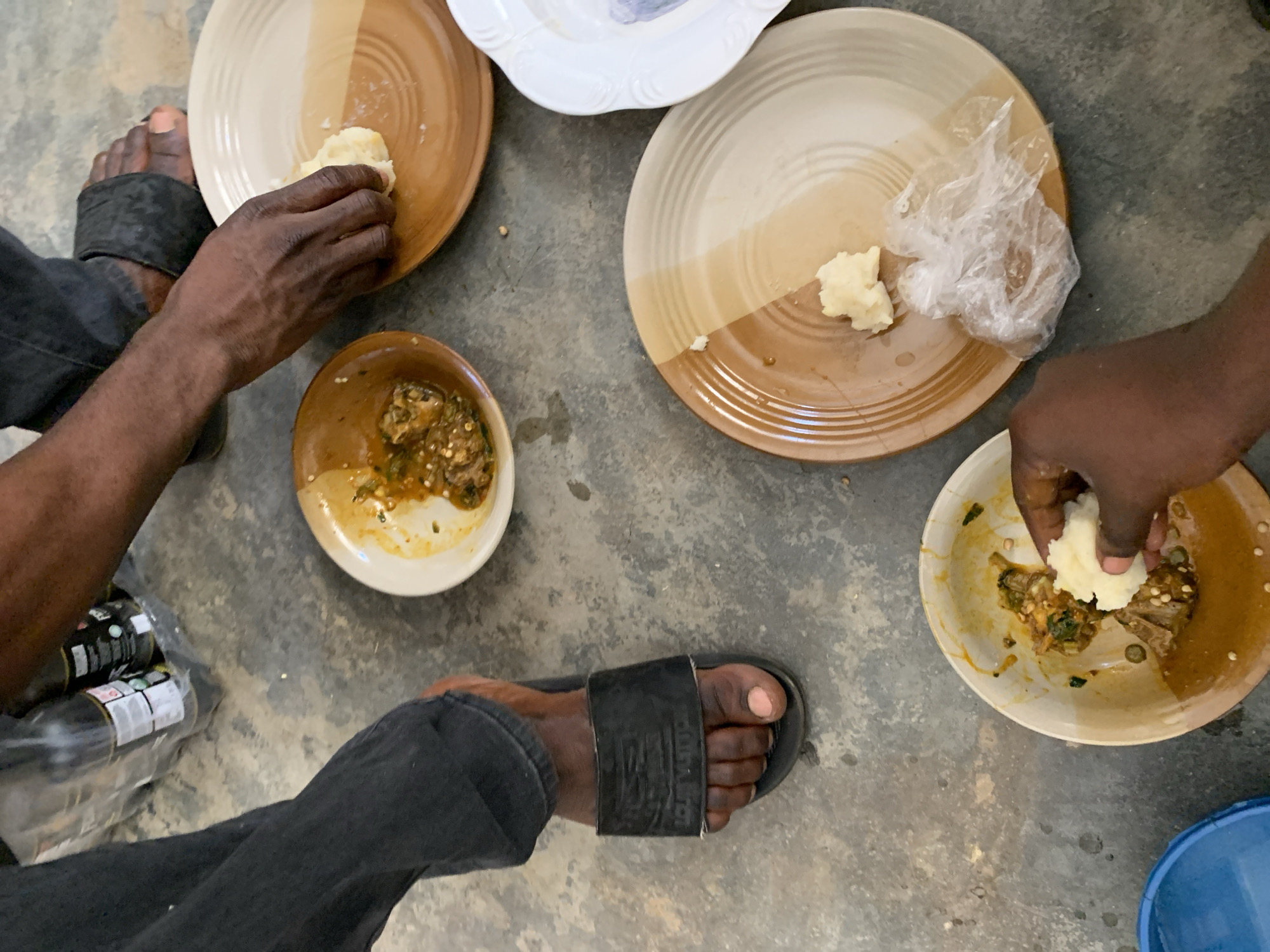 The image shows an aerial view of two people sitting on a gray concrete floor, sharing a meal. Each person is wearing dark-colored pants and sandals. One person, on the left, has two bottles in a clear plastic bag next to them. There are four plates and one small bowl arranged in a circle. Each plate contains a starchy, white, dough-like food, and one plate also has a clear plastic bag on it. The bowl contains a greenish stew. Both people are using their hands to take pieces of the dough and dip them into the stew. The location is reported to be Kabong, Nigeria, though there is no evidence to confirm this in the image.