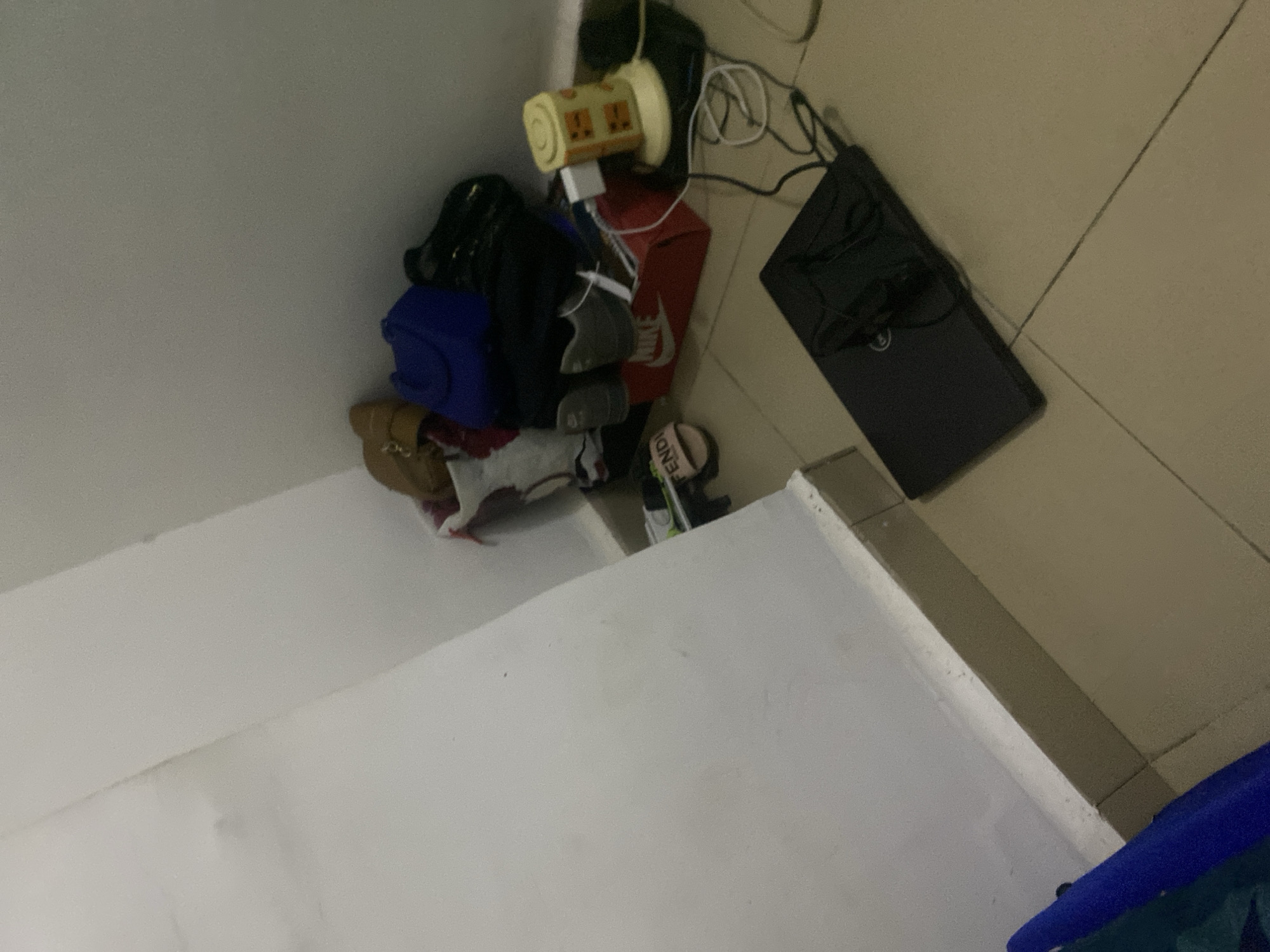 The image depicts a cluttered corner of a room. In the foreground, a black laptop is closed and placed on a beige tiled floor. Cables are connected to the laptop and extend out of view. To the right of the laptop, a yellow object, possibly a lamp or fan, with orange markings sits on a dark surface. Various items are piled in the background against a white wall, including what appears to be a red Nike shoebox, a pair of grey athletic shoes, several bags of different colors (blue, brown, and black), and a white plastic container. A pair of pink slide sandals with the word "FENDI" printed on them is visible near the floor. The environment suggests a personal living space or storage area. There is no indication of the city of Howayi, Nigeria, in the image itself.