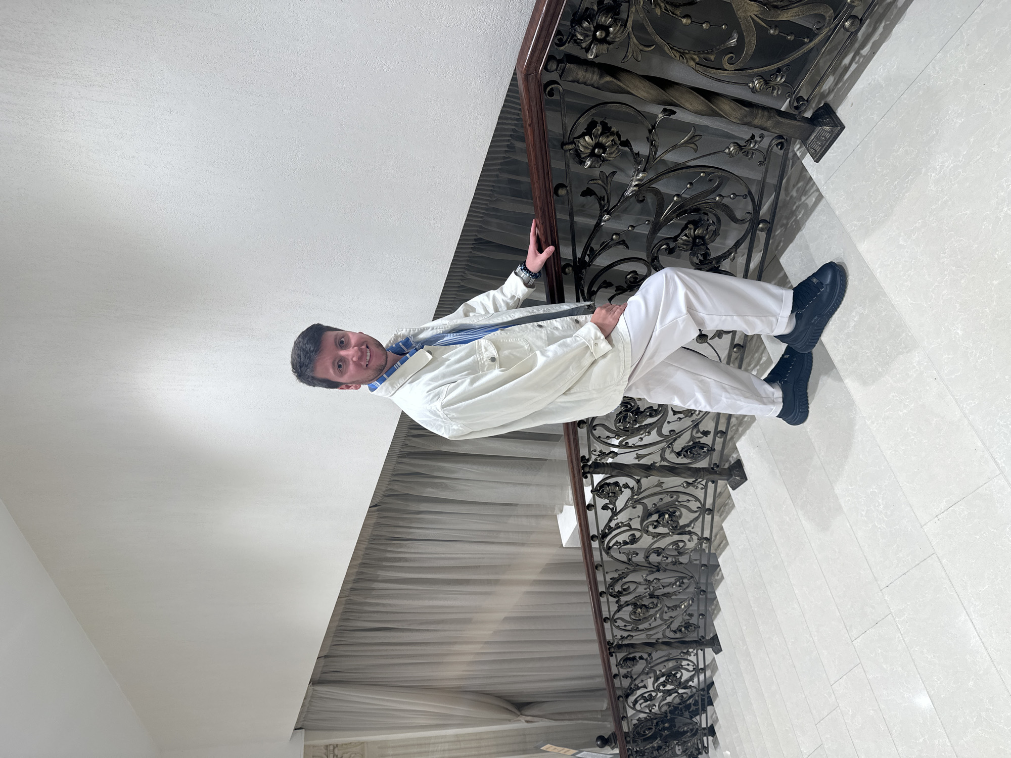This media file shows a person standing on a staircase. The person is wearing a white jacket, a blue and white striped shirt, and white pants. They are also wearing black shoes. The staircase has a decorative black metal railing with an ornate design. The railing is attached to a dark brown wooden banister. The walls are white, and the floor appears to be made of light-colored tiles. To the left of the staircase, there are white sheer curtains.
The person is smiling and looking at the camera. Their posture suggests they are casually posing for the photo. The lighting in the image is bright, indicating it is likely daytime. The overall scene appears to be indoors, possibly in a residential building or a hotel, given the style of the staircase and curtains. There is no visible text in the image. The location context provided is Moscow, Russia, which does not offer further visual cues within the image itself.