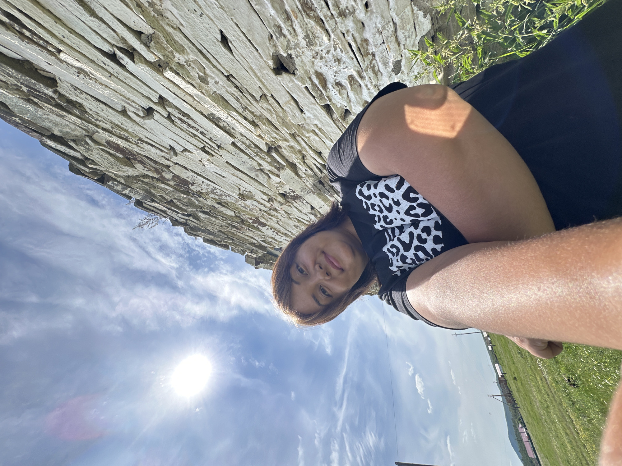 In a low-angle, outdoor selfie, a woman with shoulder-length brown hair smiles at the camera. She wears a black t-shirt with a white leopard print pattern. Her arms are crossed in front of her, and the image captures her from the chest up. Behind her, a sunlit, rough-hewn stone wall extends upwards, forming the dominant background element. To the left of the wall and in the upper portion of the frame, a bright sun shines through a partly cloudy blue sky with wispy white clouds. The lower left corner of the image shows a grassy field with some sparse yellow flowers and a distant view of rolling hills and what appear to be buildings or structures. The overall scene suggests a rural or historical setting under a bright, sunny sky.