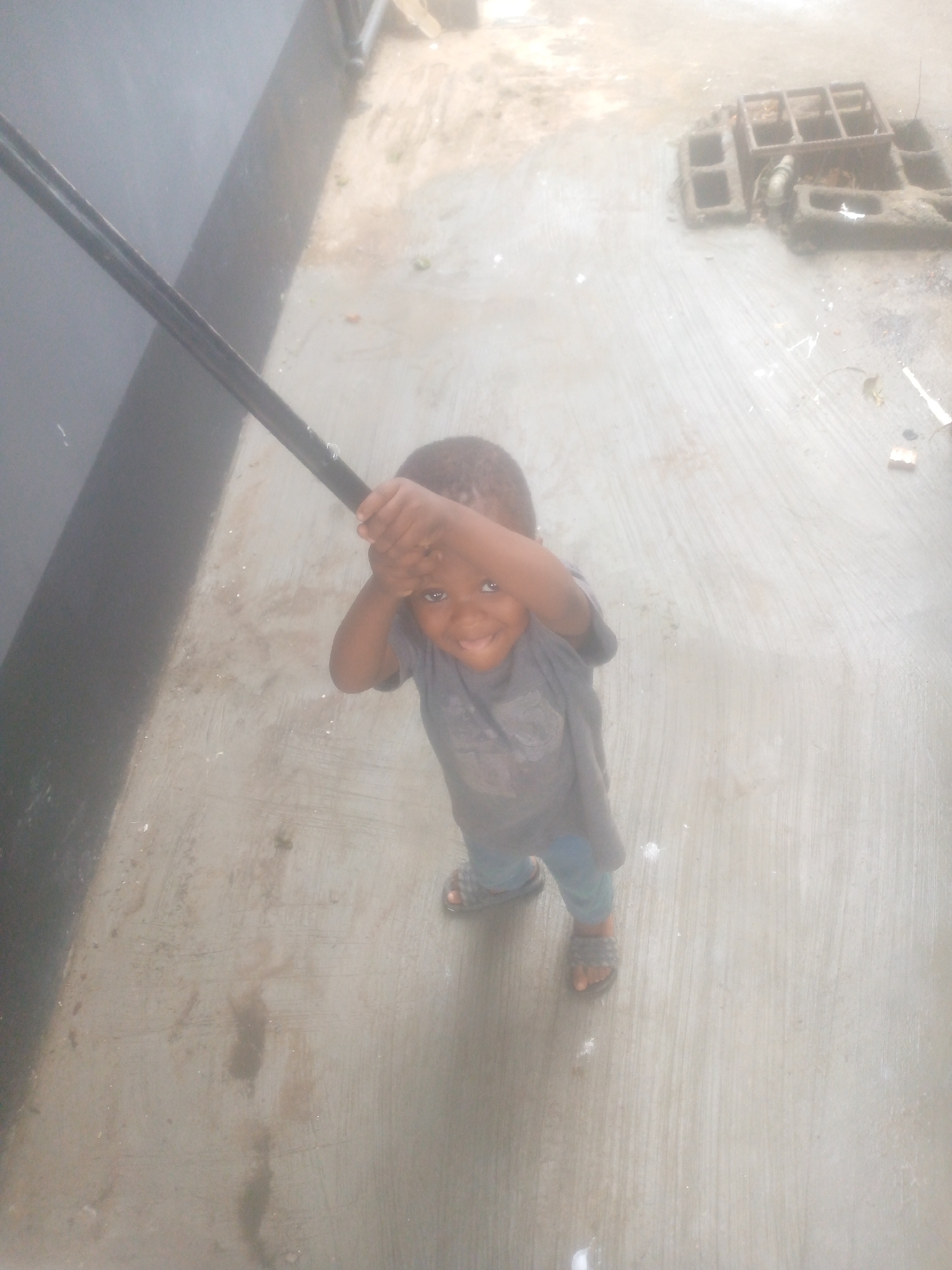 A child stands outdoors on a concrete surface. The child is holding a dark, thin, possibly metal, object above their head with both hands. The child is wearing a gray shirt, blue pants, and sandals. To the left is a dark, smooth wall. In the background to the right is a grid-like object on the concrete. The city and state are not discernible from the image.