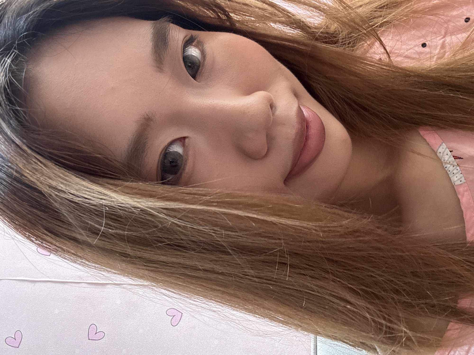 A close-up, head-and-shoulders selfie of a young Asian woman with long, straight, light brown hair is shown. She has light brown eyes, light makeup, and is wearing a pink top with a subtle pattern. The background is a pink wall with small, light pink heart designs. The image appears to have been taken indoors, likely in a residential setting. The woman is looking towards the camera.