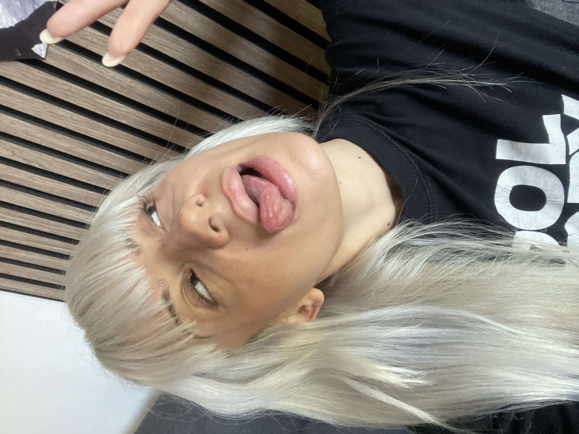 A woman with platinum blonde hair is lying on her back, sticking her tongue out and looking up. She is wearing a black shirt with white lettering. Her fingers are visible in the upper left corner. The background includes a textured wall or headboard with horizontal stripes. The location is unknown, and there is no indication that it is in Tilburg, Netherlands.