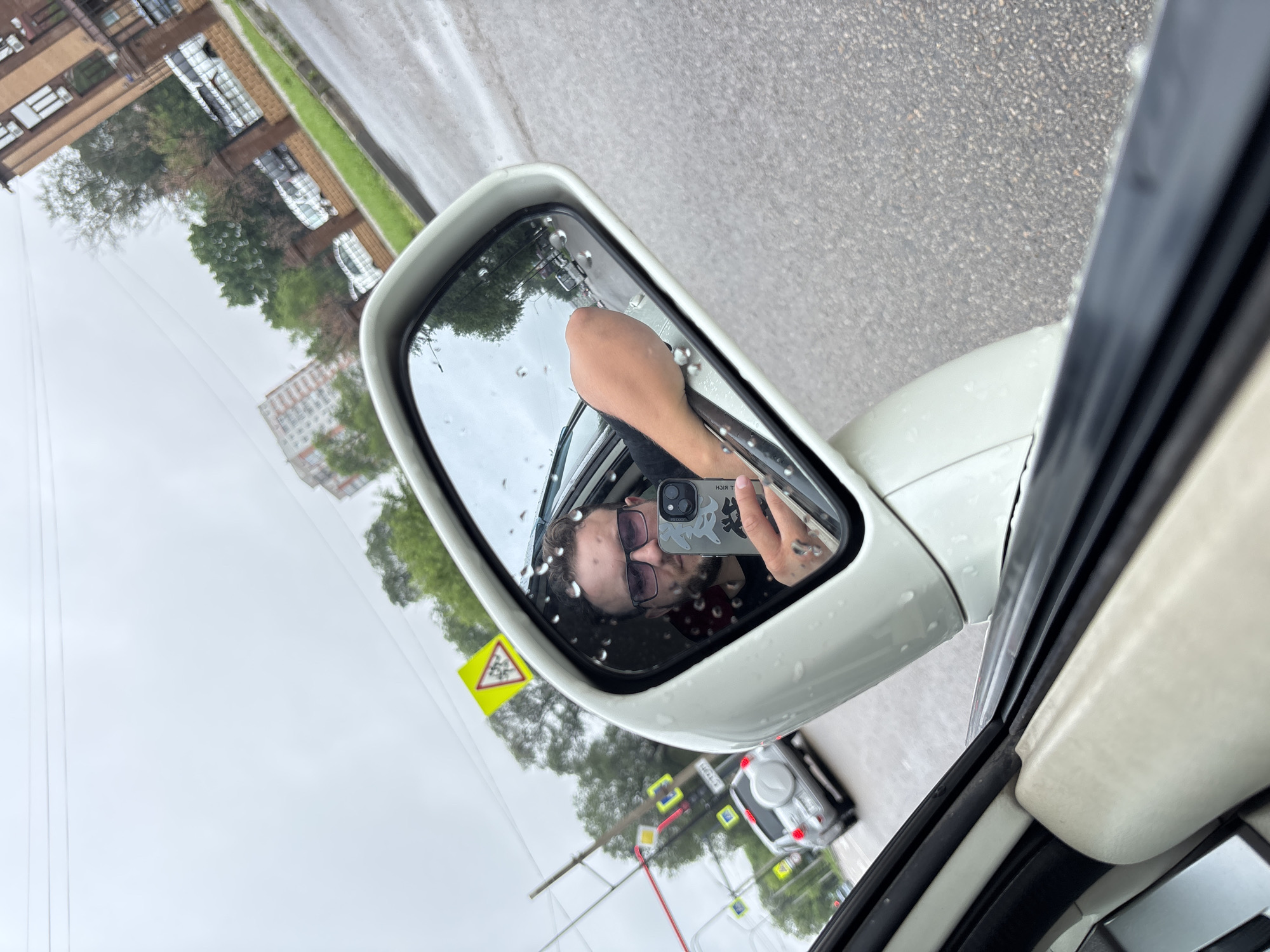 A man takes a selfie in the side-view mirror of a car. He is wearing sunglasses and holding a smartphone. The car is on a street in Blagoveshchensk, Russia, on a rainy day. In the background, there are buildings, trees, and other cars. The street is wet, and there are raindrops on the mirror.