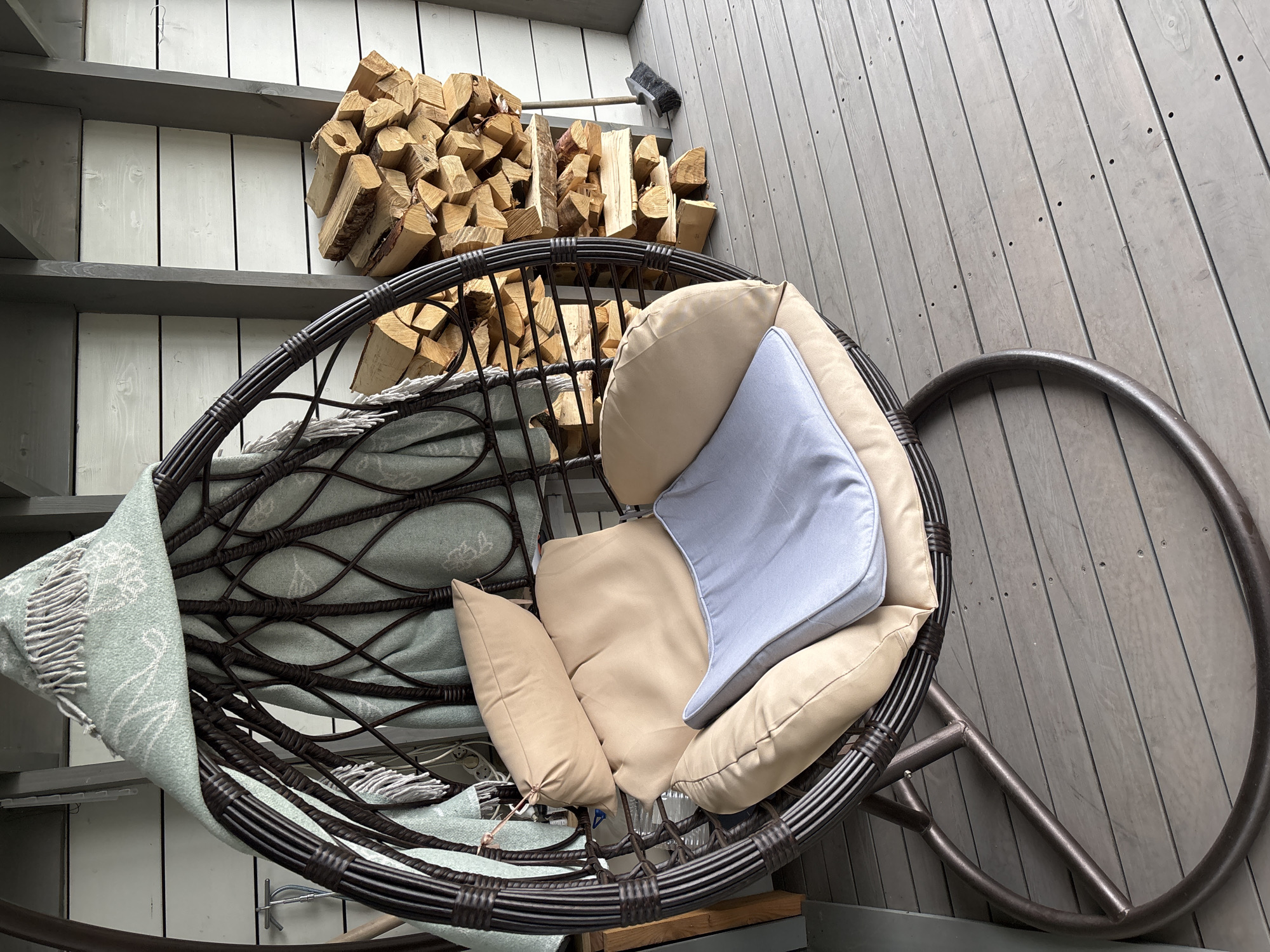 The image depicts an outdoor patio, likely in a residential setting, as evidenced by the presence of stacked firewood and a hanging chair. The patio is constructed of weathered grey wooden planks, laid horizontally. To the right, a substantial pile of cut firewood is neatly stacked against a white wooden wall, which is further segmented by darker grey vertical beams. A simple broom with a dark head and wooden handle rests against the firewood.
Dominating the left and center of the frame is a large, dark brown wicker hanging chair, designed for relaxation. It is suspended by a metal frame and equipped with plush cushions in two shades: a light tan and a pale blue. A light green throw blanket, featuring a subtle pattern and fringed edges, is draped over the top of the chair.
The background shows a portion of a building with horizontal white siding and vertical grey structural beams. The overall impression is one of a rustic, comfortable outdoor space. There are no people visible in the image. The lighting suggests it is daytime, likely overcast. The specific location in Zvenigorod, Russia, is not discernible from the visual elements present.