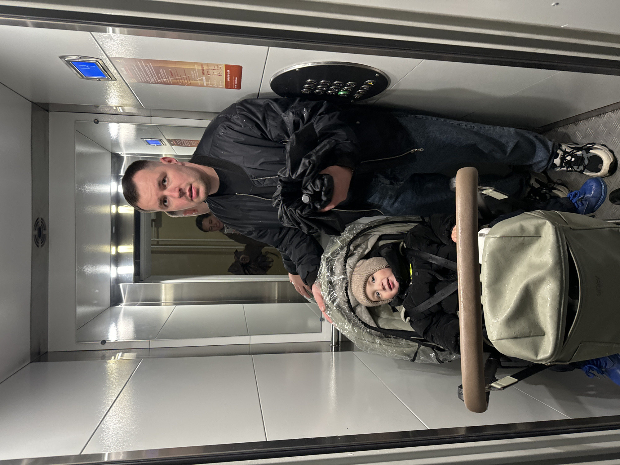 A man and a baby in a stroller are inside an elevator. The man is wearing a black jacket and blue jeans, and the baby is wearing a beige beanie. The stroller has a rain cover. The elevator has a mirrored back wall and a control panel with buttons. The location is Akulovo (Gp Kubinka), Russia.