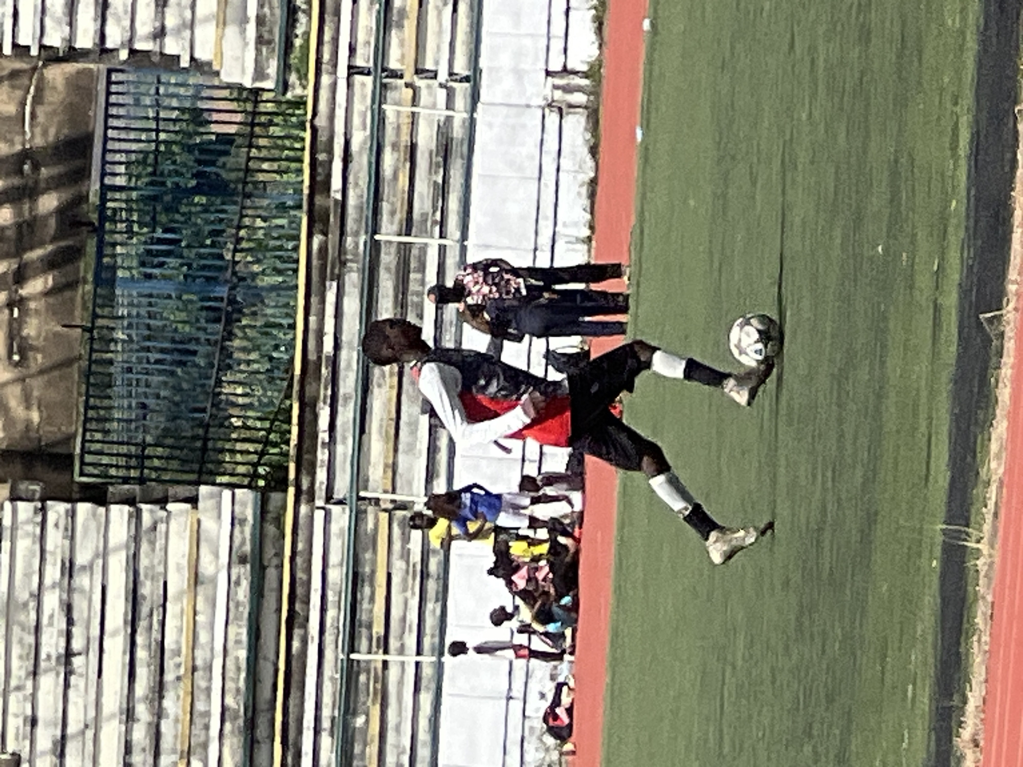 A male soccer player is running with a soccer ball on a green field in New Stadium, Nigeria. He is wearing a red and white jersey and black shorts. There are several spectators in the background, some sitting and some standing. The stadium appears to be empty except for the player and the spectators. The sky is clear and the sun is shining.
