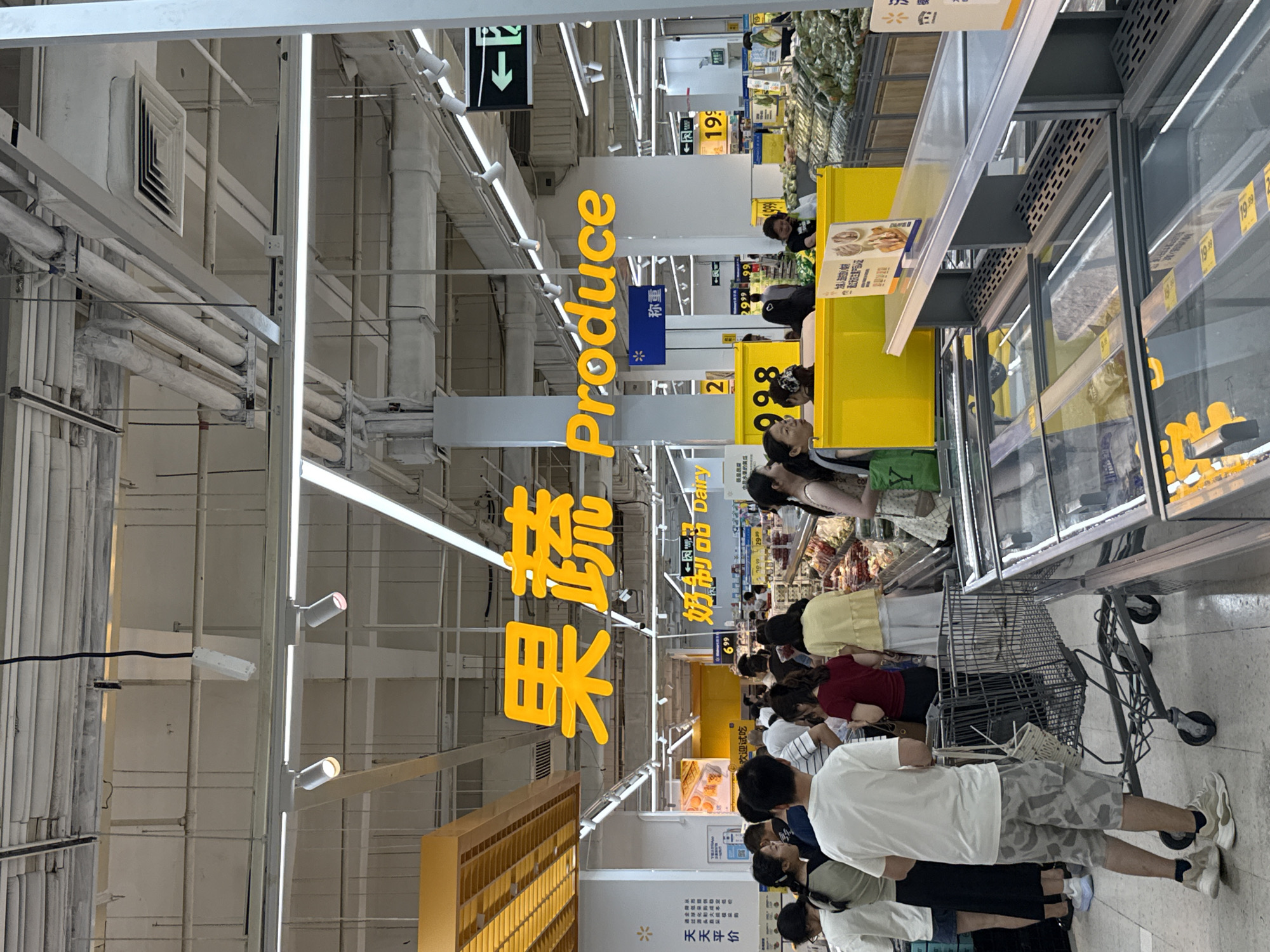 A wide-angle, eye-level shot shows a bright, indoor supermarket aisle in Huizhou, China. A large, bright yellow sign in Mandarin characters "果蔬" and English "Produce" is prominently displayed overhead, indicating the produce section. Below the sign, a crowd of shoppers, predominantly Chinese, are navigating the aisle. Several individuals are pushing shopping carts, while others are browsing or conversing. In the foreground, a man in camouflage shorts and a white t-shirt stands with his shopping cart. To the right, a glass-fronted refrigerated display case is visible, stocked with produce. The ceiling of the supermarket features exposed beams, lighting fixtures, and ventilation systems, typical of a large retail environment. Signs with prices and product information are visible throughout the aisle, and a blue Walmart logo is present on a sign. The overall atmosphere appears to be a busy, functional supermarket setting.