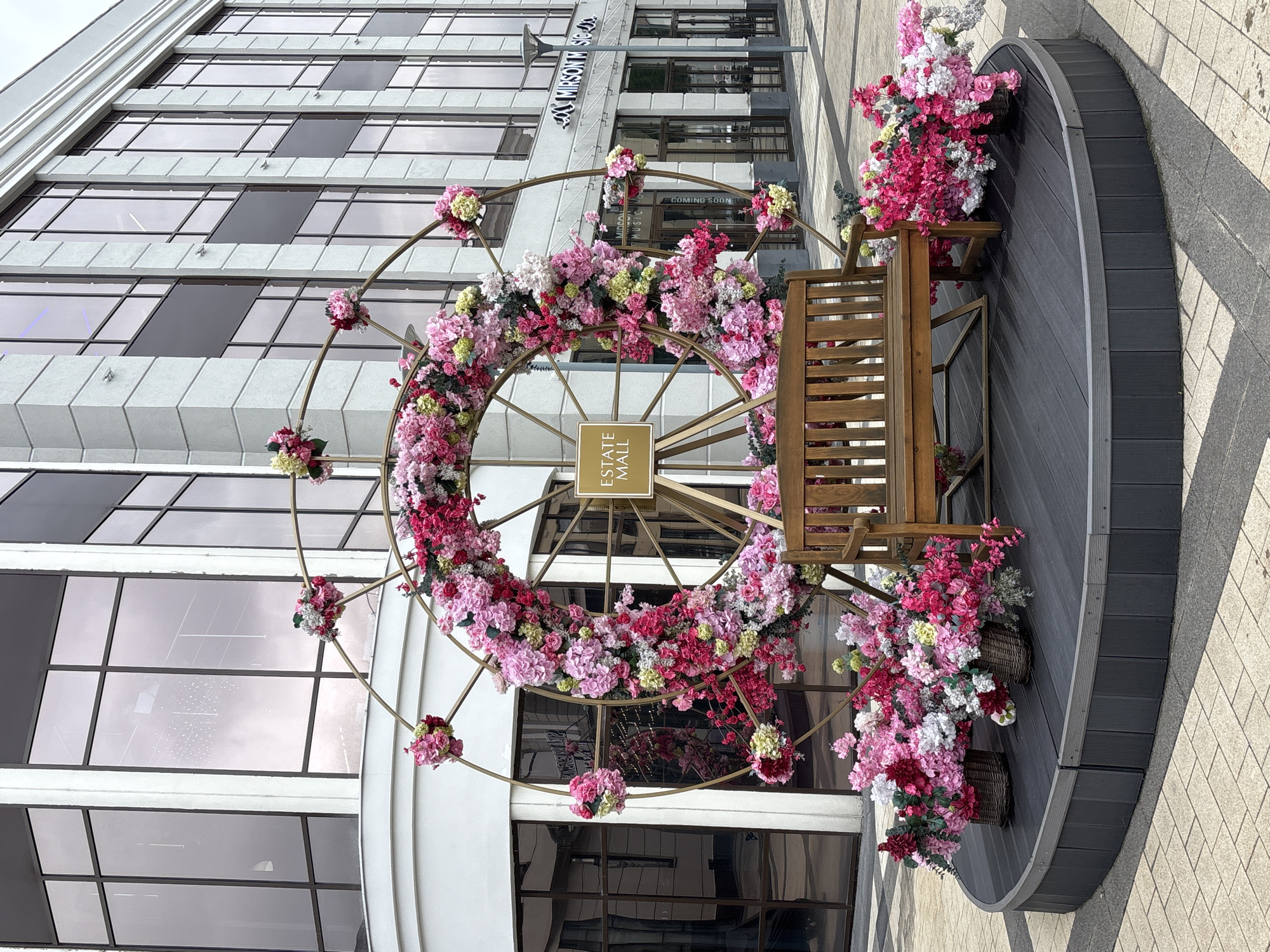 A large, circular floral installation decorated with pink and white flowers sits on a raised wooden platform. A wooden bench is placed in front of the installation. In the background, a modern building with large windows is visible. A sign on the installation reads "ESTATE MALL". The scene appears to be outdoors in a city. The image does not contain any people or specific identifiable landmarks for the city of Monolit, Russia.